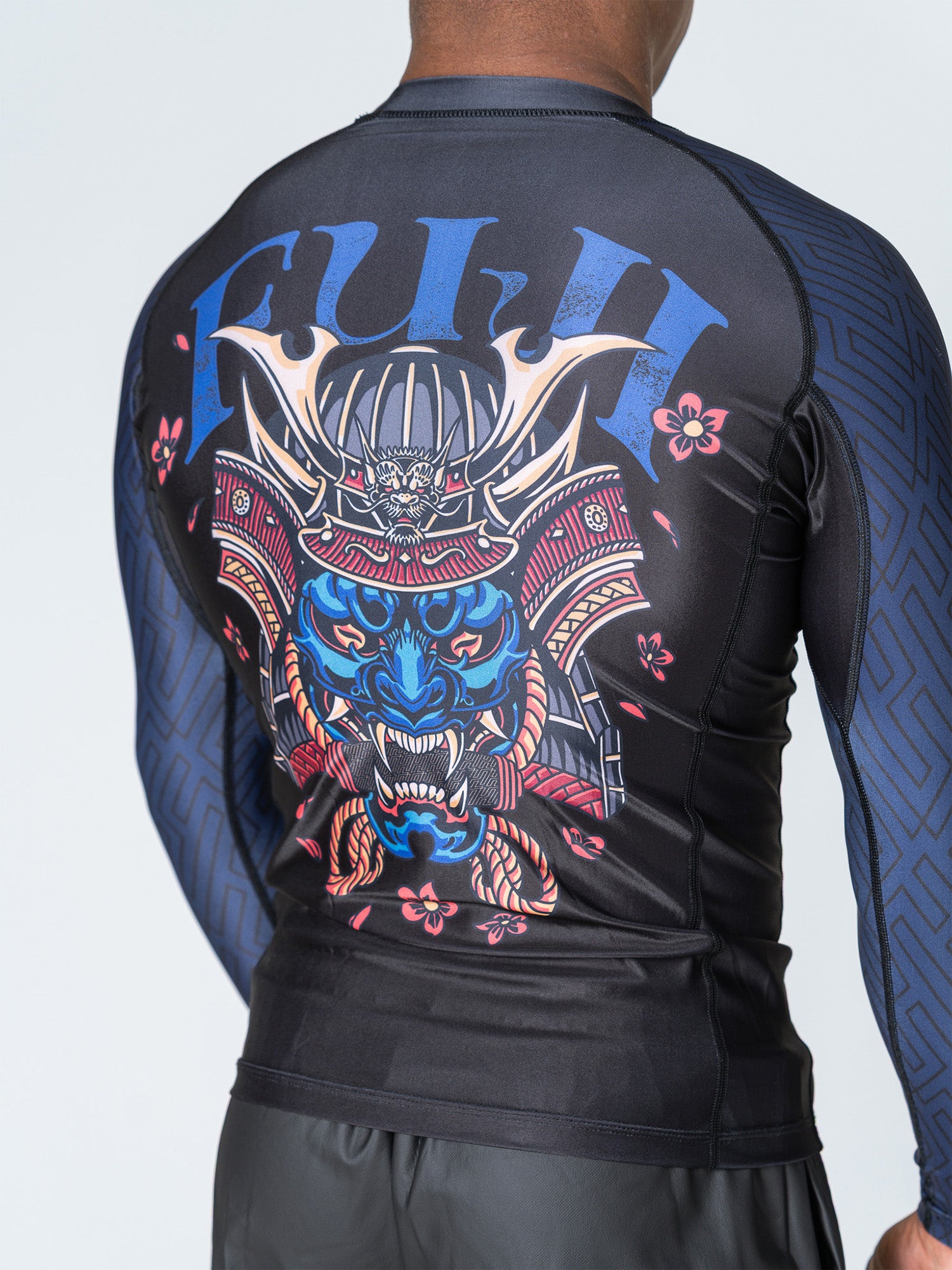 A person wearing the Oni Samurai Flex Lite Rashguard Blue, a black long-sleeve with blue geometric sleeves and a vibrant back design featuring a samurai helmet, tiger face, flowers, and bold blue "FUJI" lettering.