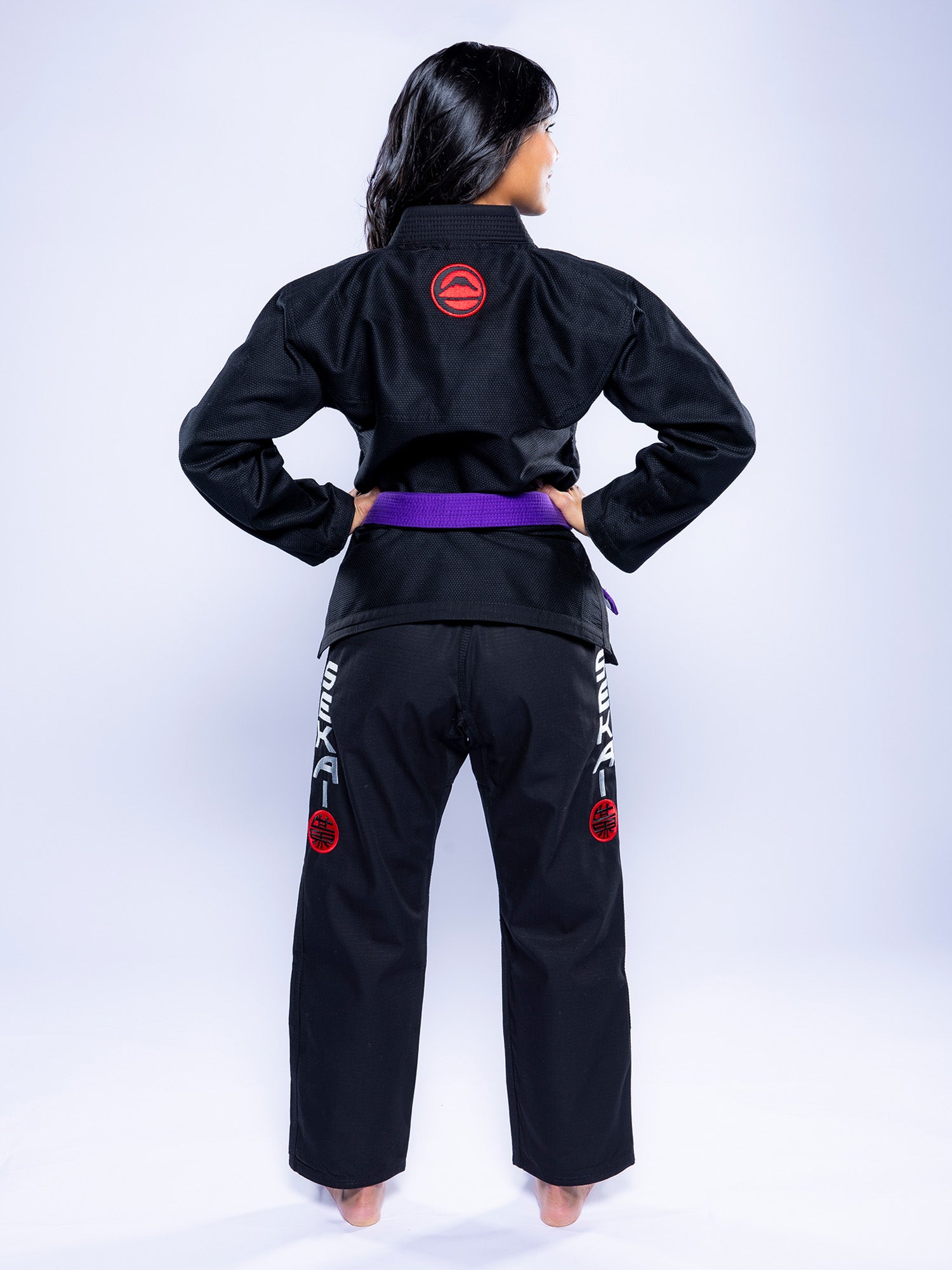 A person with long dark hair stands barefoot, facing away, wearing the Womens SEKAI 3.0 BJJ Gi Black Grey adorned with a purple belt. Red and white patches are seen on the back and legs of the uniform.