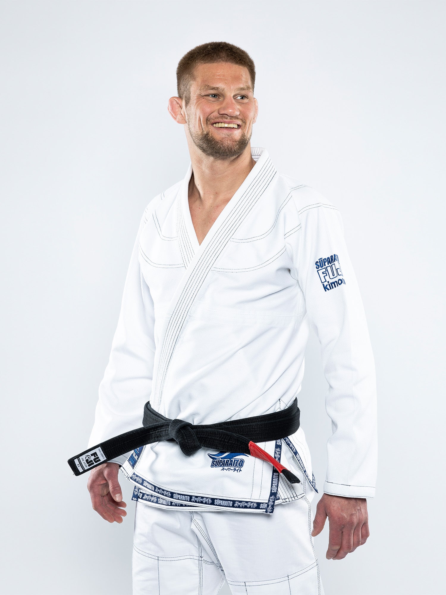 A man wearing the Suparaito BJJ Gi with a black belt stands smiling and looking to his left against a plain white background.