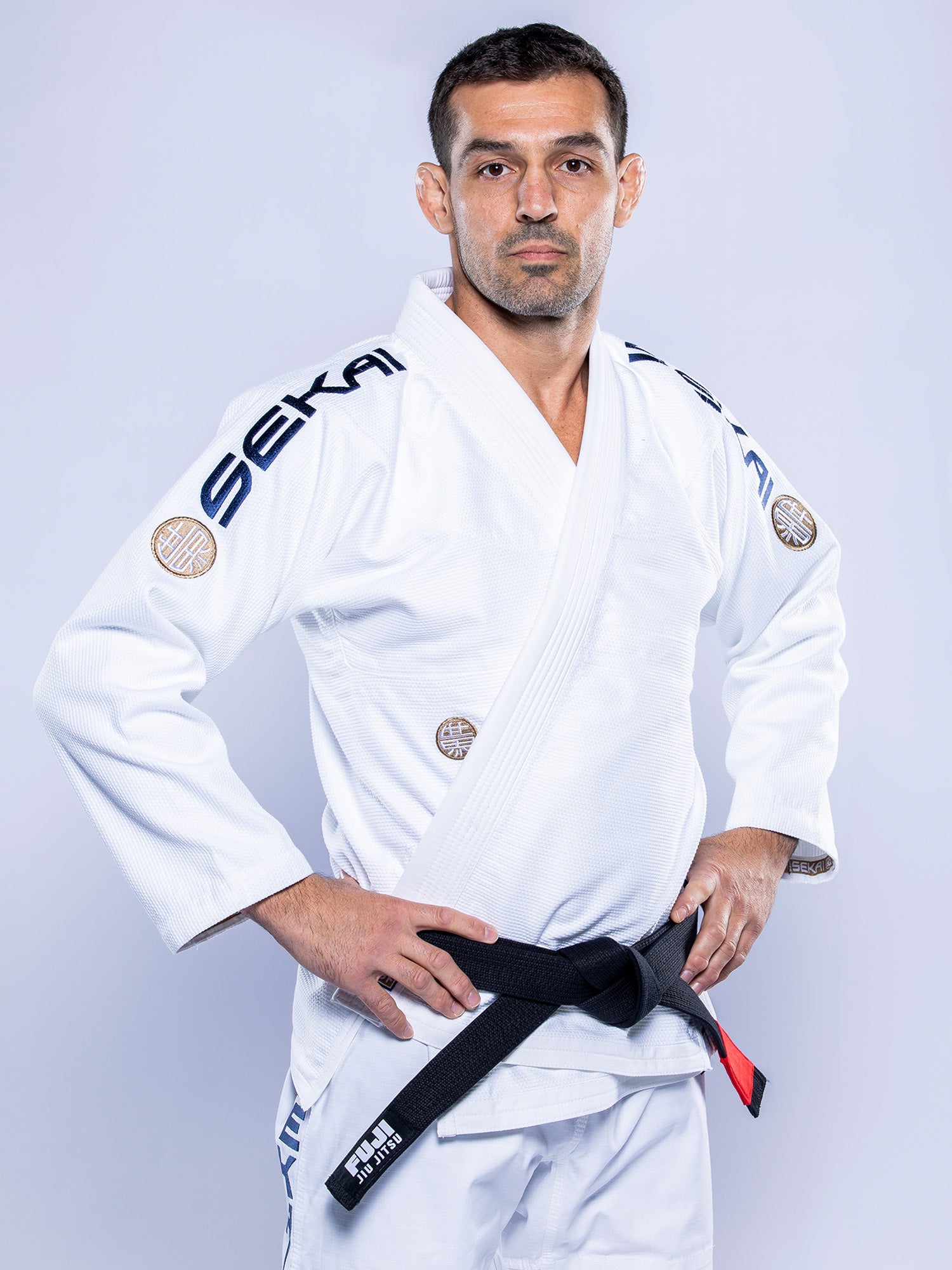 A man with short dark hair and a black belt stands hands on hips in a white SEKAI 3.0 BJJ Gi (White Navy), featuring "SEKAI" on the shoulders, against a light gray background. He has a serious expression.
