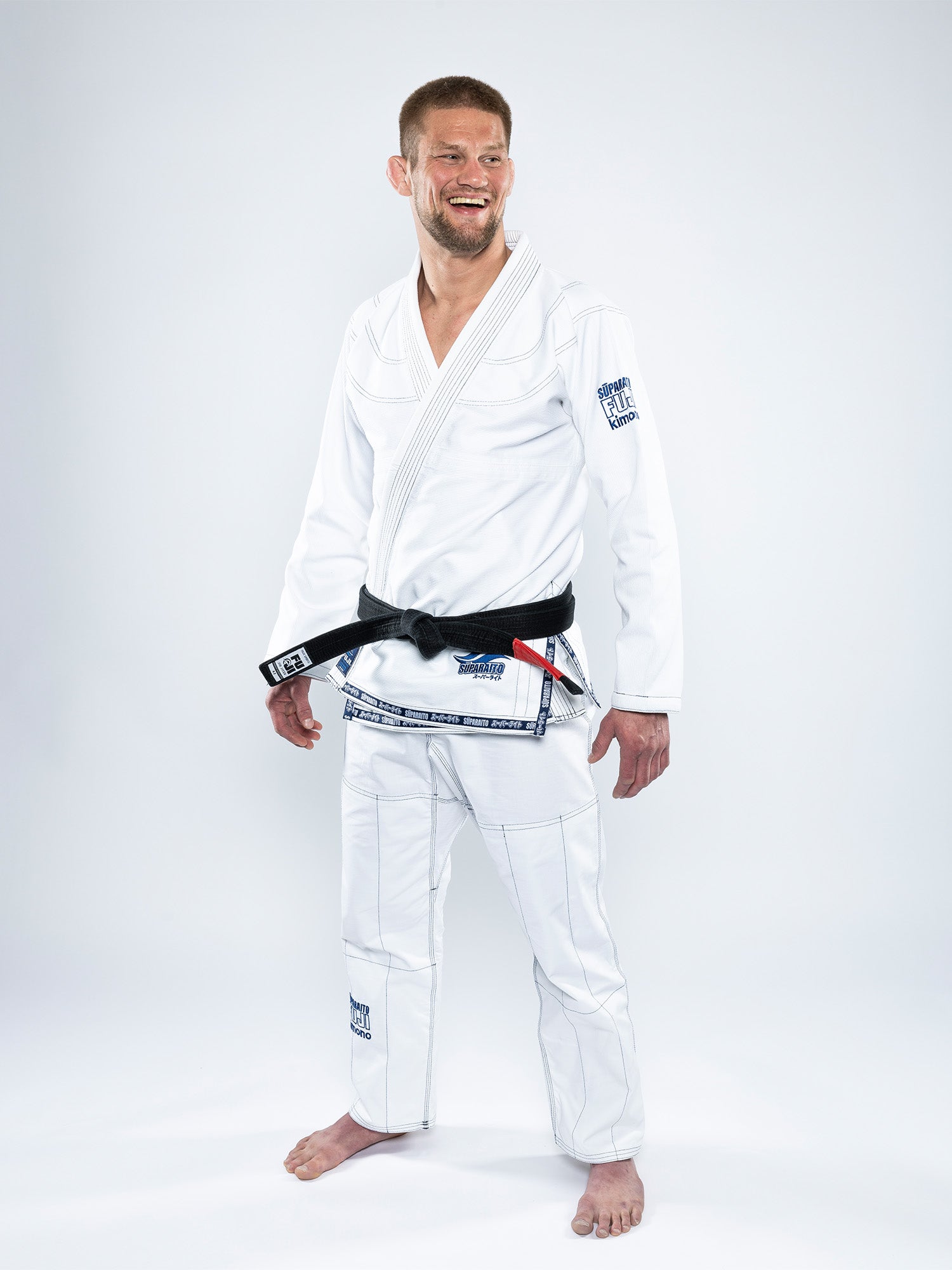 A man wearing the Suparaito BJJ Gi with a black belt stands barefoot on a white background, smiling and glancing slightly to the side.