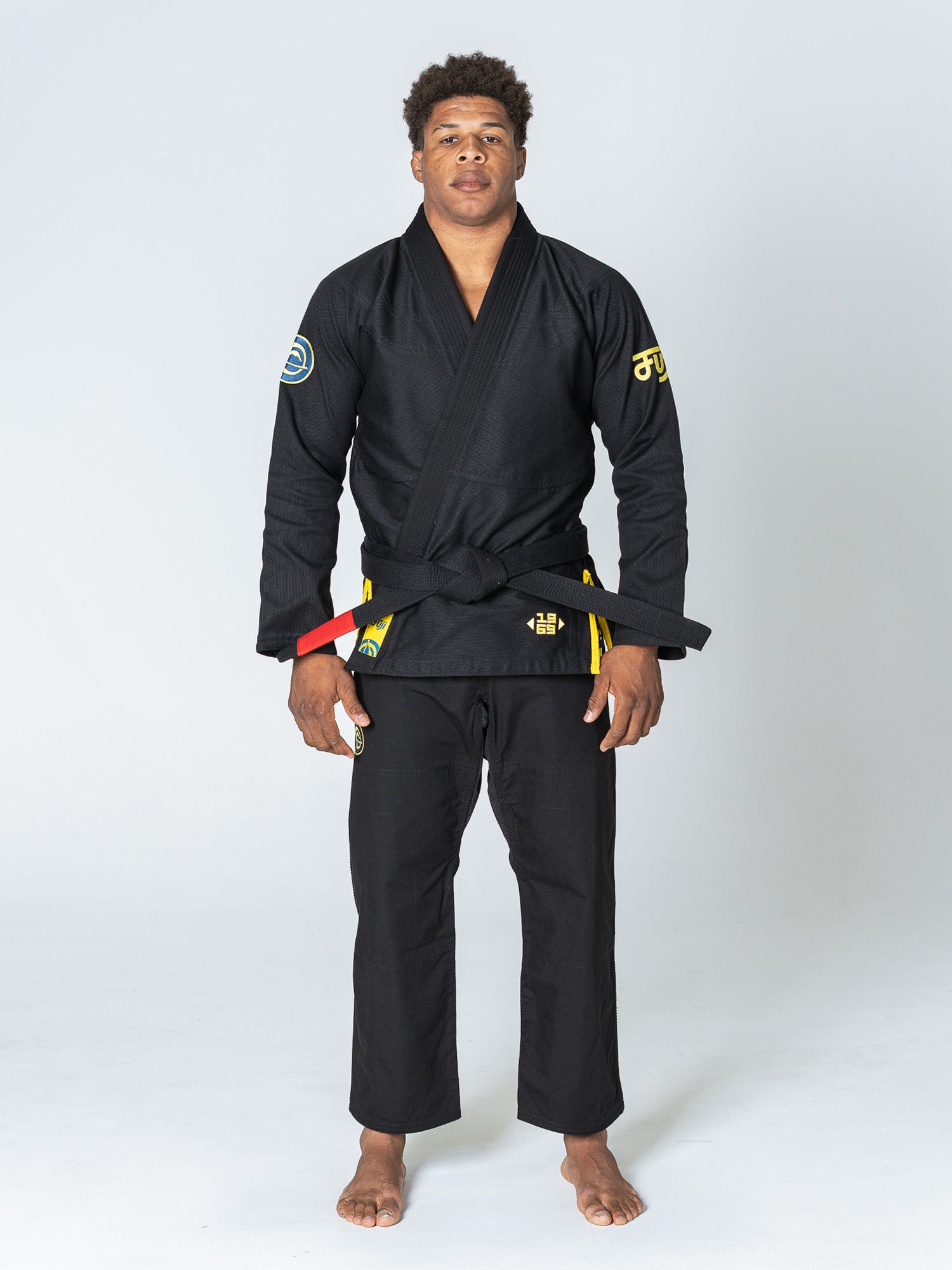 A man stands barefoot on a white background wearing the Special Edition Holiday Flow Tech Jiu Jitsu Gi Black with patches and a black belt tied around his waist.