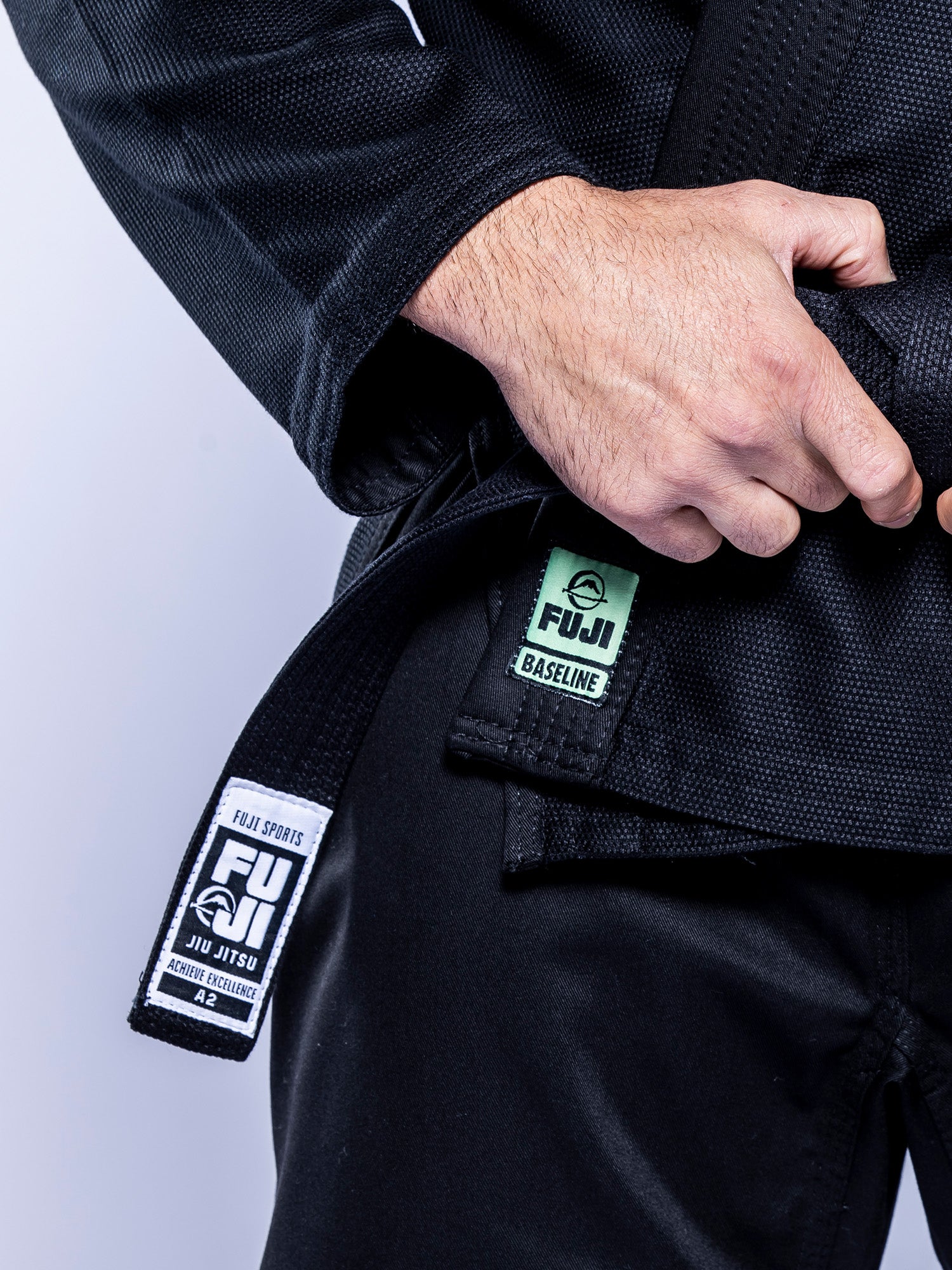 A person wearing a Baseline 2.0 BJJ Gi Black Mint grips the lapel. The gi displays branded patches and a black belt with a white Fuji Sports label.