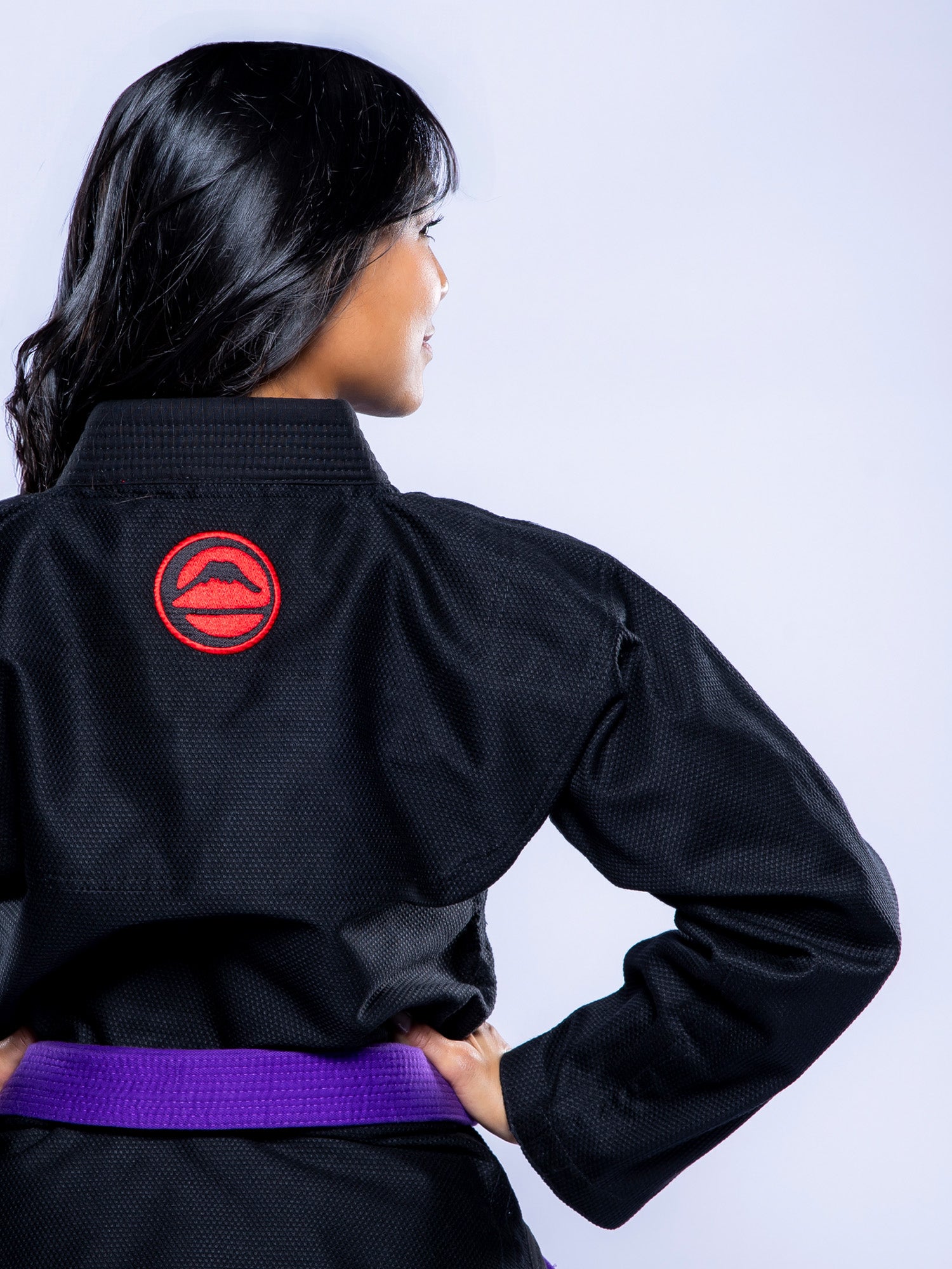 A woman with long dark hair stands facing away from the camera, hands on hips, wearing the Womens SEKAI 3.0 BJJ Gi Black Grey and a purple belt, against a light background.