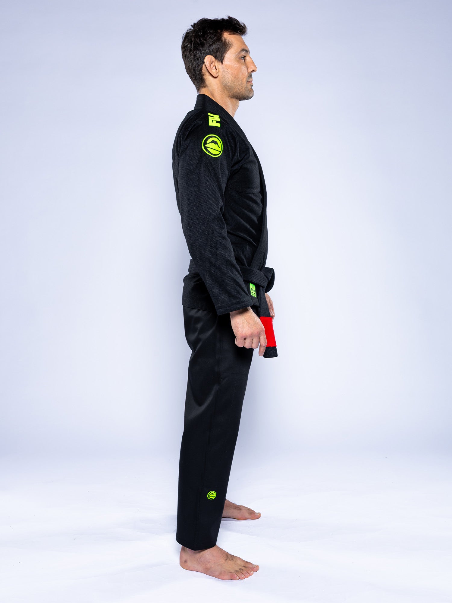 A man wearing the Baseline 2.0 Slim Fit BJJ Gi Black Green stands barefoot in side profile against a plain light background, holding a red and black belt at his waist.