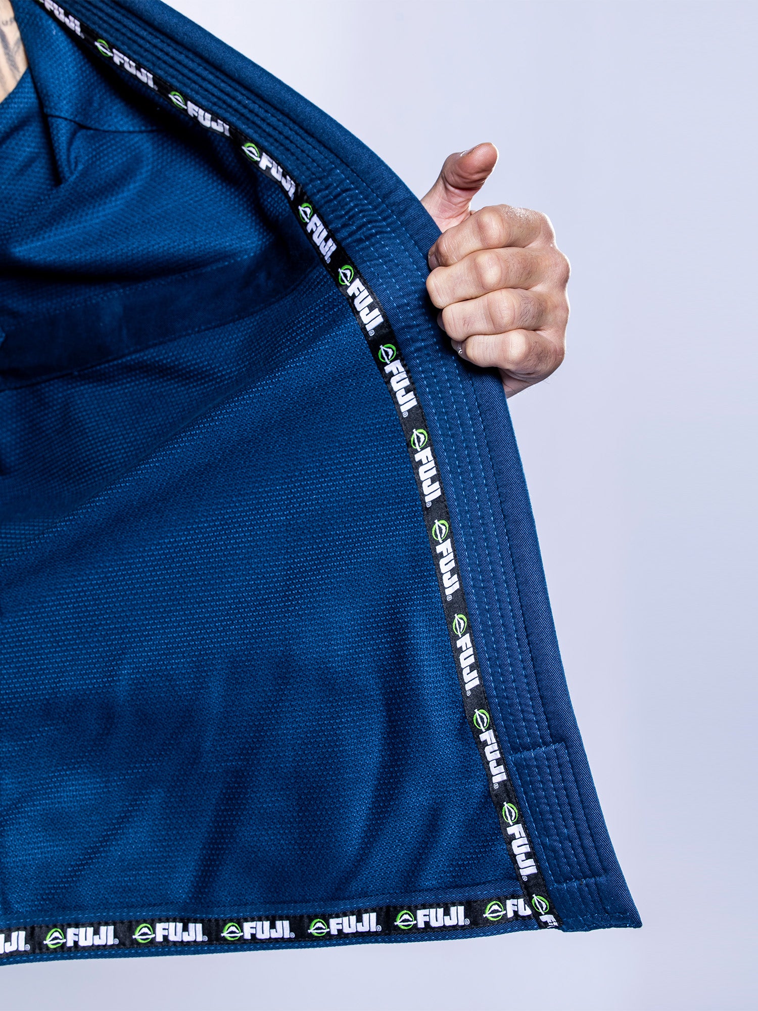A person opens the lapel of the Lightweight BJJ Gi Navy, showing its inner lining and a trim featuring repeated FUJI logos in white and green. The background is plain and light-colored.