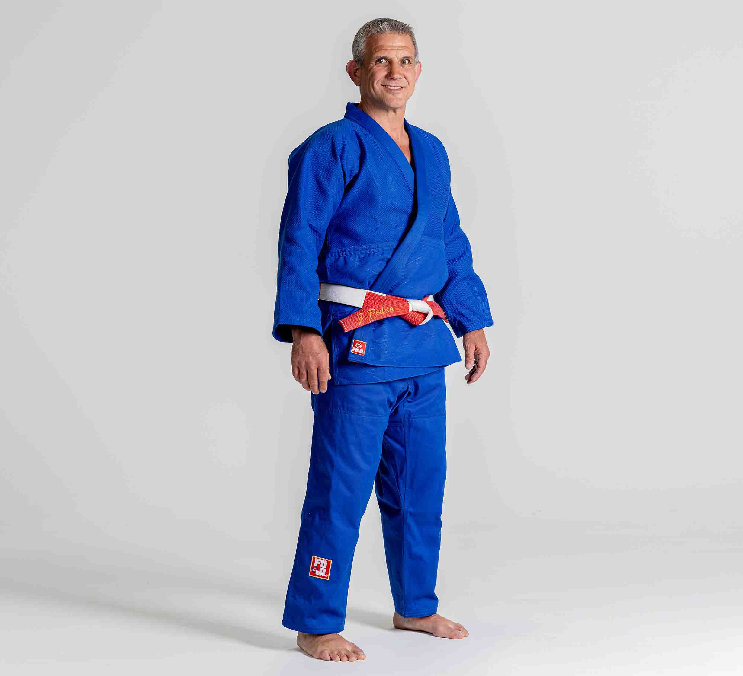 A man wearing the Euro Competition Judo Gi Blue and a red-and-white belt stands barefoot on a plain light background, smiling at the camera.