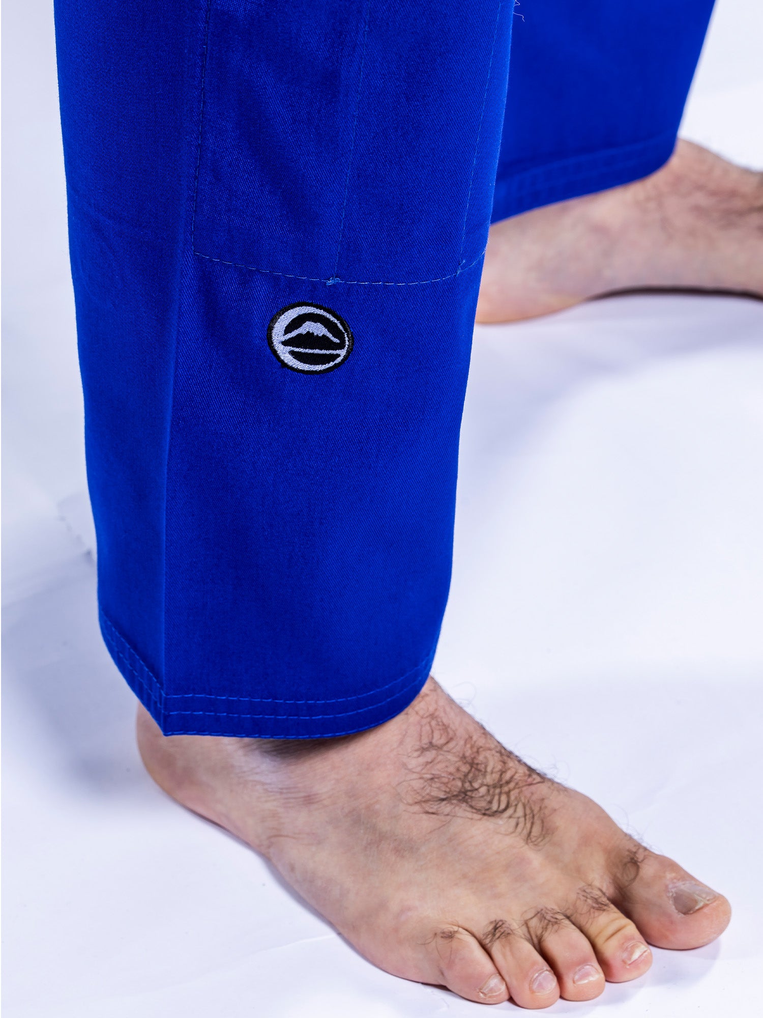 Close-up of a person's lower legs and bare feet in the Baseline 2.0 BJJ Gi Blue White, featuring bright blue pants with a small circular logo near the hem, standing on a white surface.