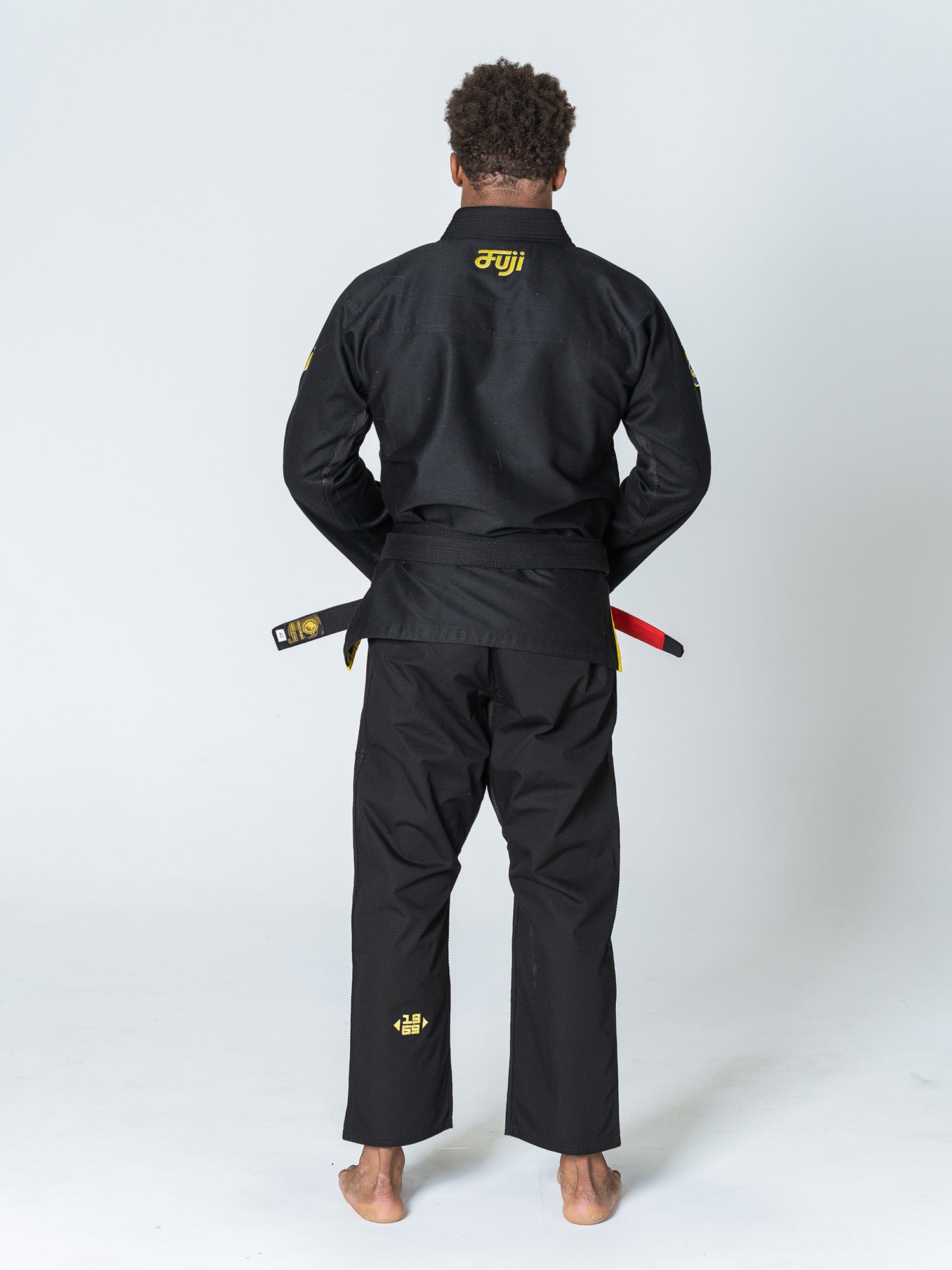 A person stands barefoot facing away, wearing the Special Edition Holiday Flow Tech Jiu Jitsu Gi Black with yellow embroidery and a black belt with a red bar, against a plain white background.