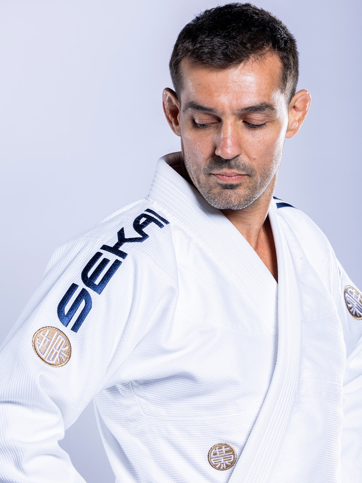 A man wearing the SEKAI 3.0 BJJ Gi White Navy, featuring embroidered patches and SEKA on the shoulder, looks down seriously against a plain light background.