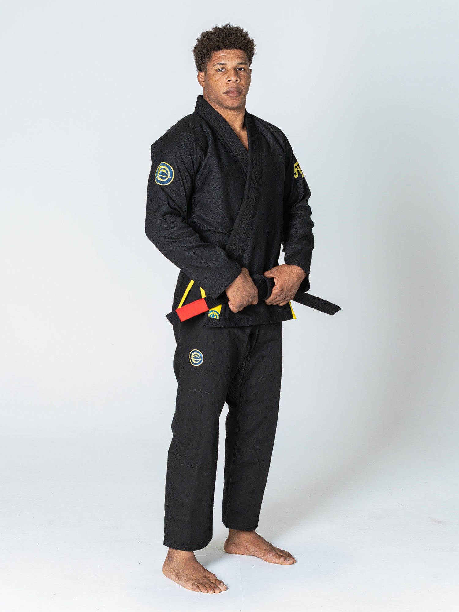A man wearing the Special Edition Holiday Flow Tech Jiu Jitsu Gi Black with yellow trim stands barefoot on a white background, holding a black belt with a red bar.
