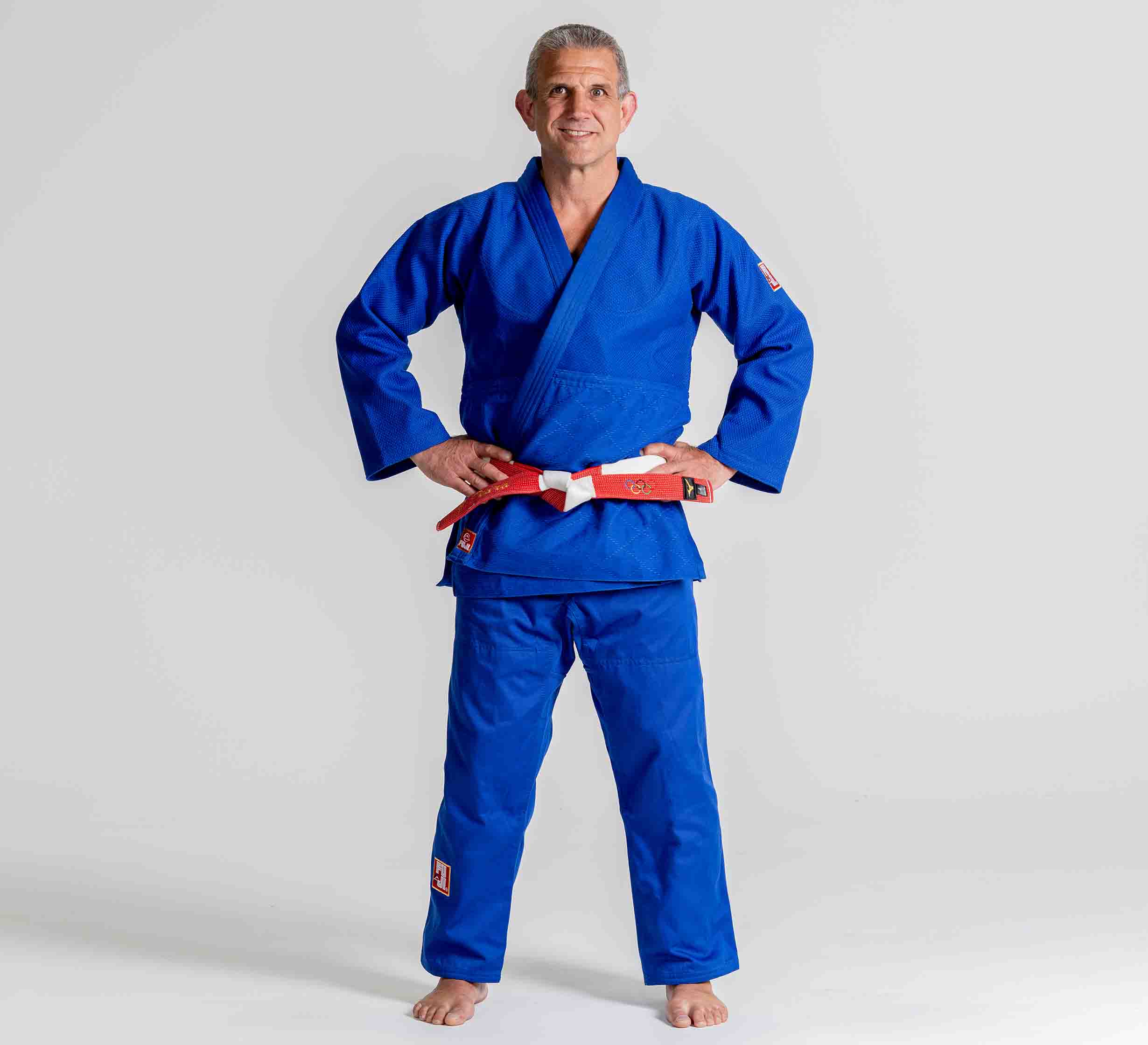 A man stands barefoot on a plain background, smiling with his hands on his hips while wearing the Euro Competition Judo Gi Blue with a red and white belt.