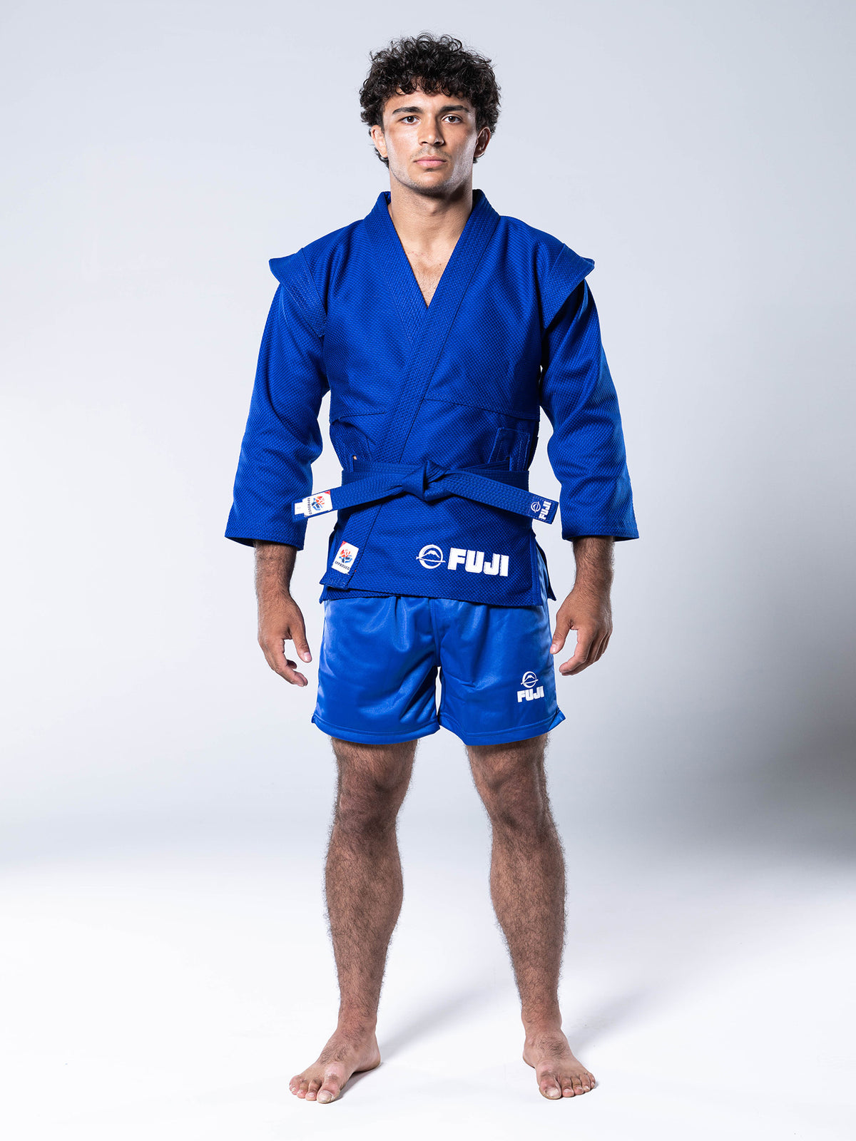 A man stands barefoot in a blue martial arts gi jacket and FIAS Sambo Shorts Blue with FUJI logos, against a plain light background.