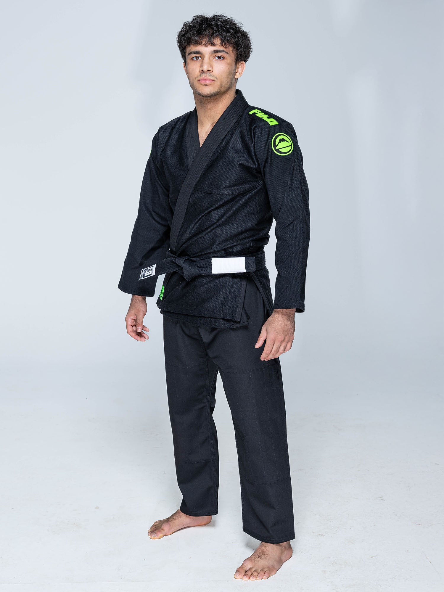 A person stands barefoot on a white floor, wearing the Baseline 2.0 Slim Fit BJJ Gi Black Green with green shoulder patches and a black belt. The background is plain and light-colored.