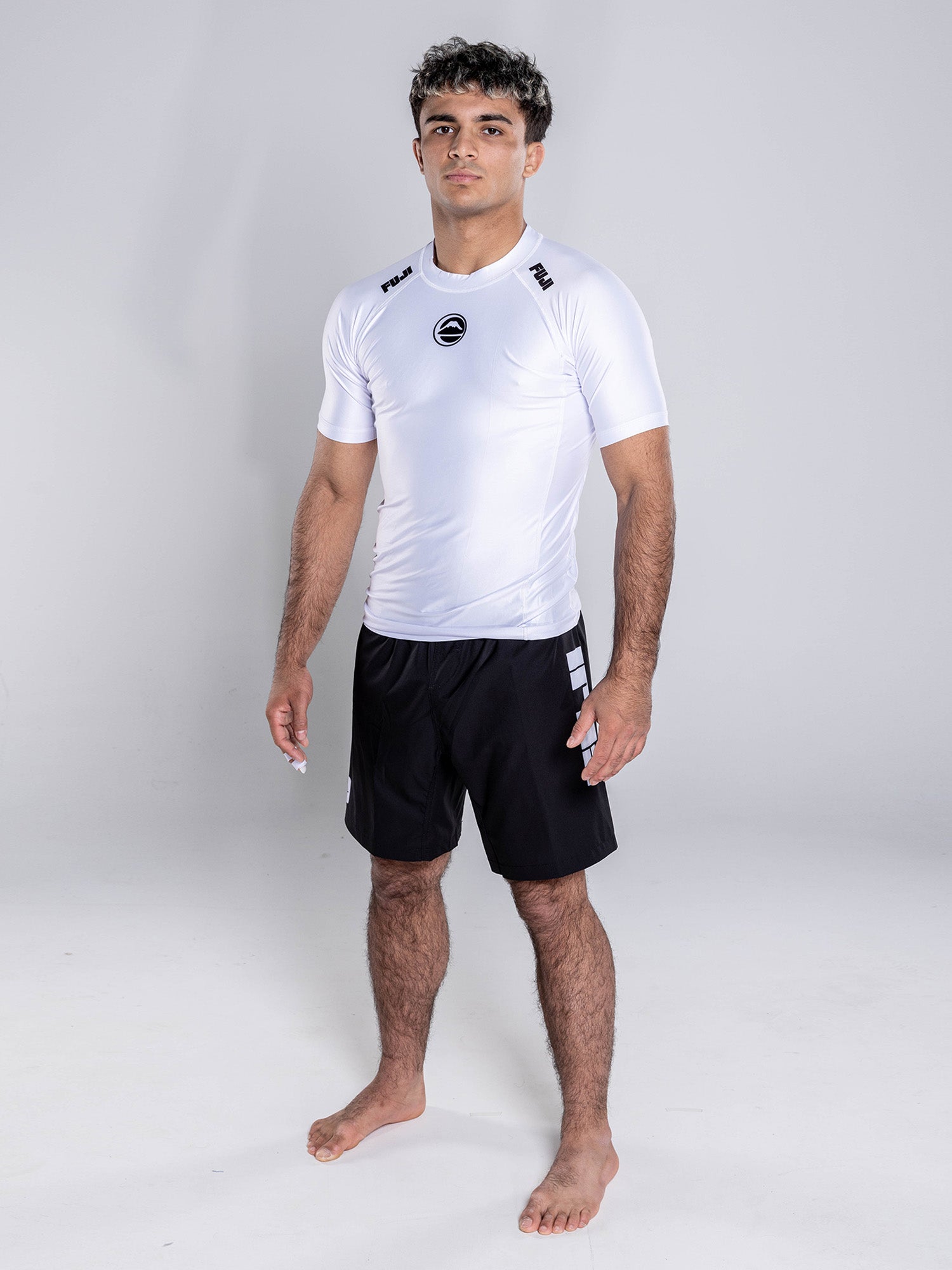 A young man with short, curly hair stands barefoot on a white backdrop, wearing the Baseline 2.0 Short Sleeve Rashguard White and black shorts with gray stripes. He faces forward, arms relaxed and expression neutral.
