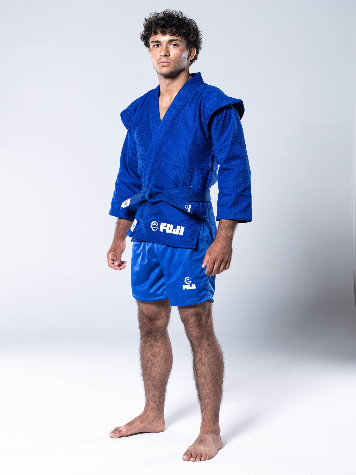 A person stands barefoot on a white background, wearing the FIAS Sambo Kurtka Jacket Blue with a matching blue belt and shorts. The jacket features FUJI branding, and the individual looks directly at the camera.