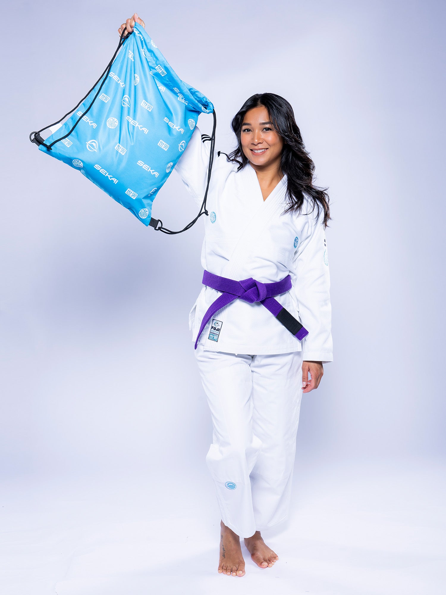 A woman in the Womens SEKAI 3.0 BJJ Gi White Teal and a purple belt stands barefoot, smiling while holding a light blue drawstring bag with white patterns against a plain light background.