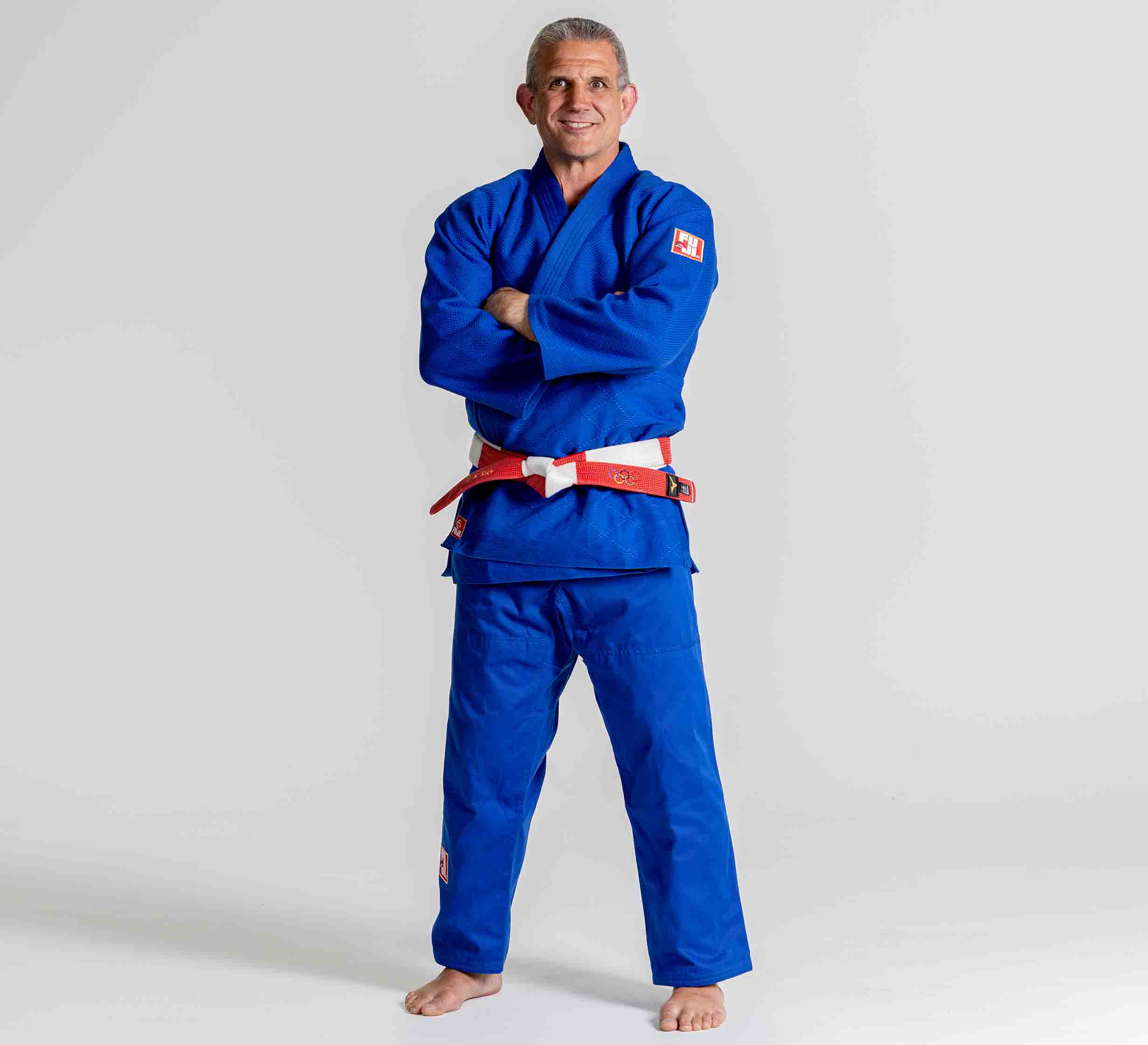 A man stands barefoot on a plain background, smiling with his arms crossed and wearing the Euro Competition Judo Gi Blue with a red and white belt.
