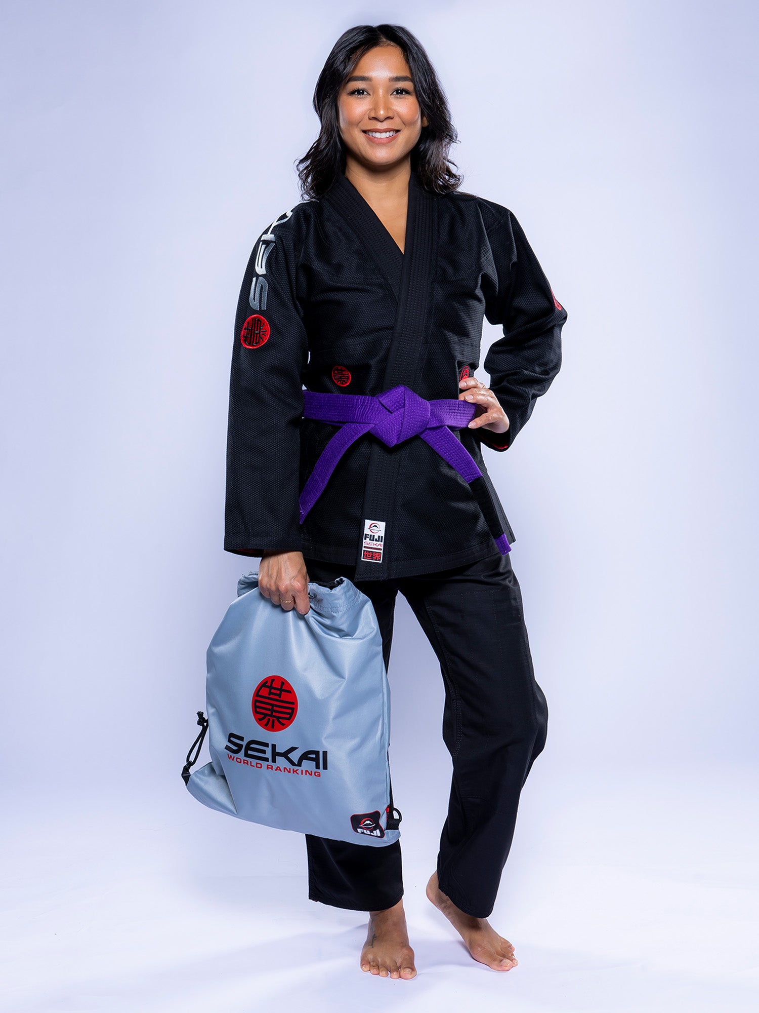 A woman in the Womens SEKAI 3.0 BJJ Gi Black Grey with a purple belt smiles barefoot, holding a light gray drawstring bag featuring red and black SEKAI branding against a plain white background.