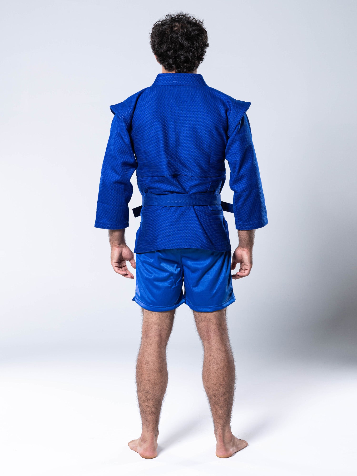A person stands barefoot facing away, wearing the FIAS Sambo Kurtka Jacket Blue with Blue Belt and blue shorts. The background is plain light gray.
