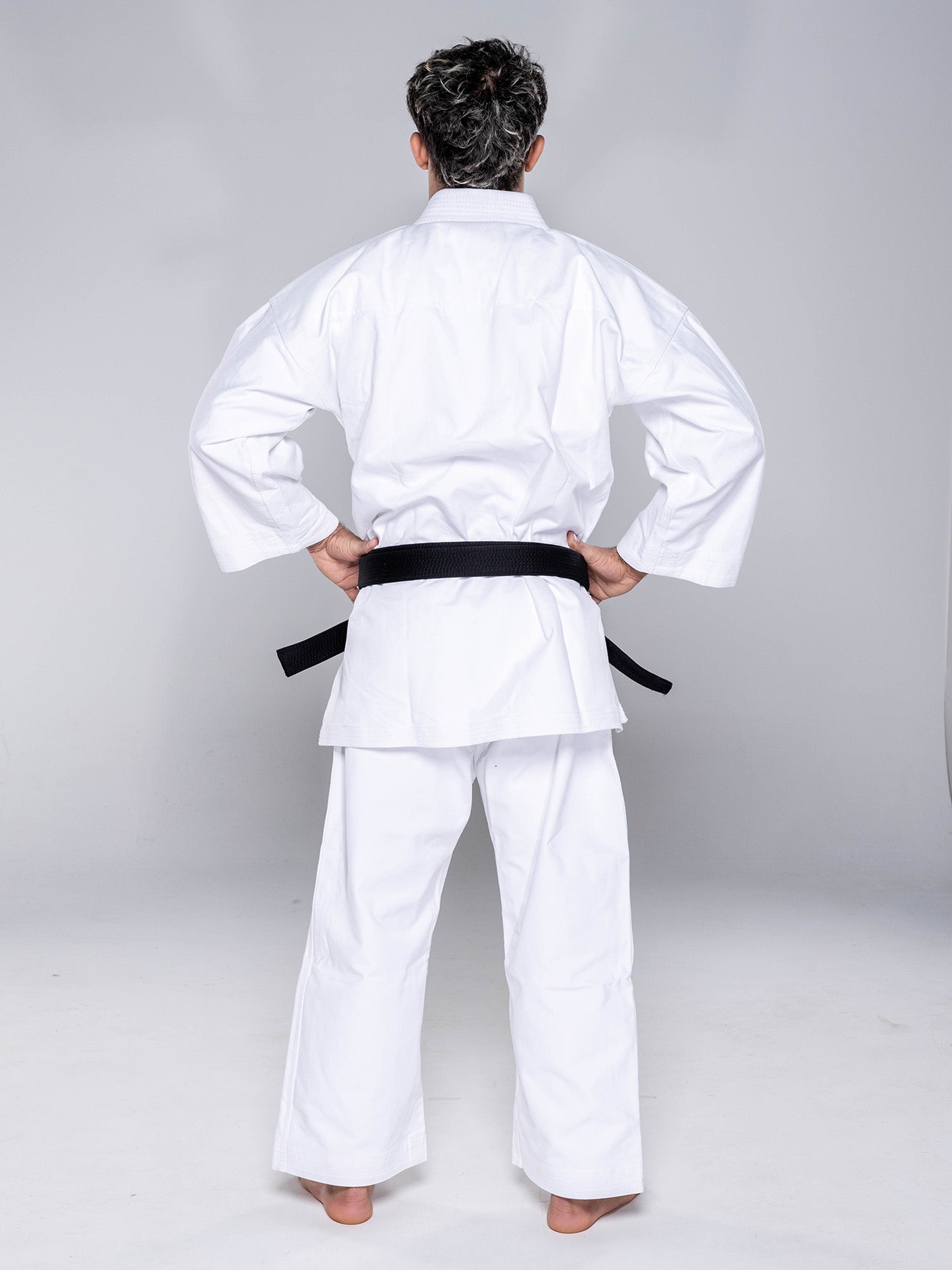 A person wearing the Kaizen Karate Gi Advanced White with a black belt stands barefoot, facing away from the camera with hands on hips, against a plain light background.