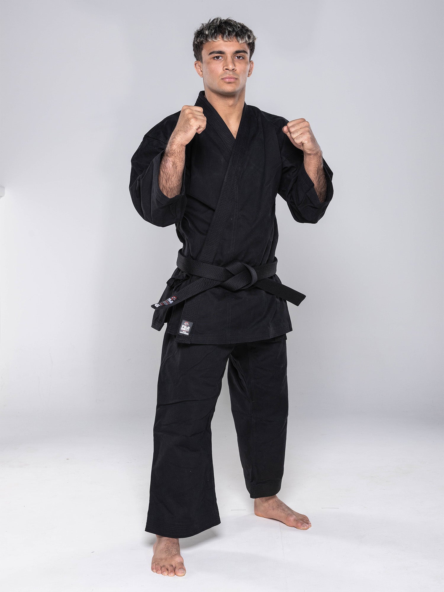Wearing the Kaizen Karate Gi Advanced Black and a black belt, a person stands barefoot on a white floor with fists raised in a fighting stance against a light background.