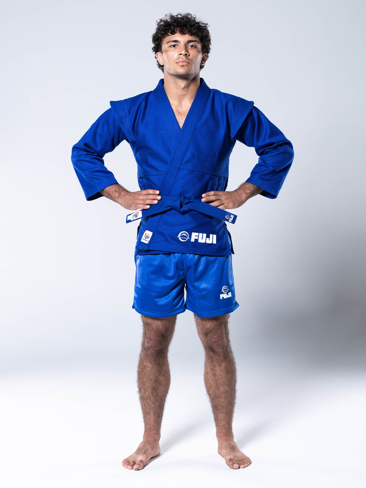 A fit, curly-haired male athlete stands against a white background wearing the FIAS Sambo Kurtka Jacket Blue with Blue Belt—a blue Fuji brand martial arts uniform featuring a long-sleeved jacket, matching blue shorts, and a blue belt—his hands placed firmly on his hips.