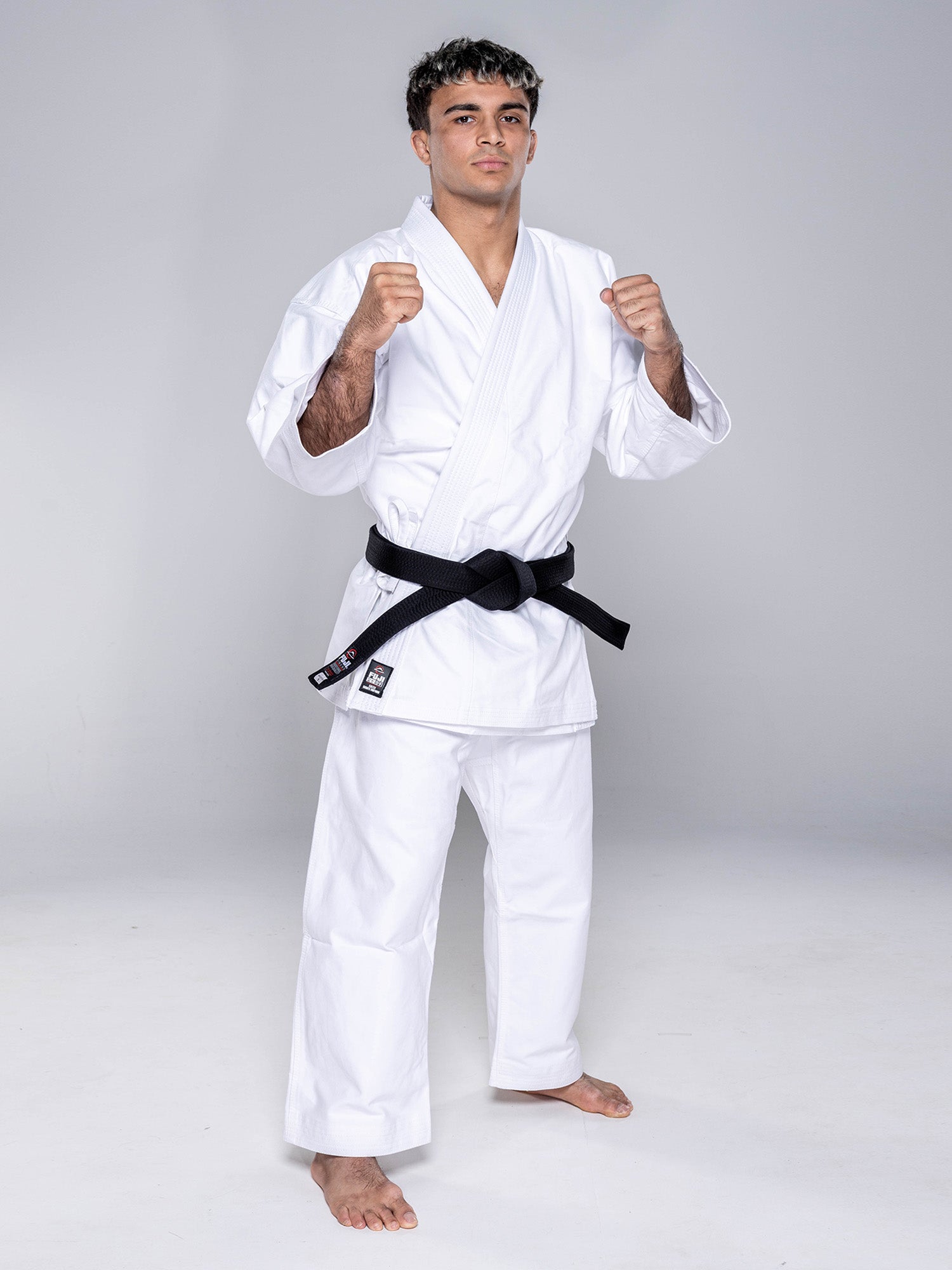 A young man stands barefoot on a plain background in a ready stance, wearing the Kaizen Karate Gi Advanced White and a black belt.