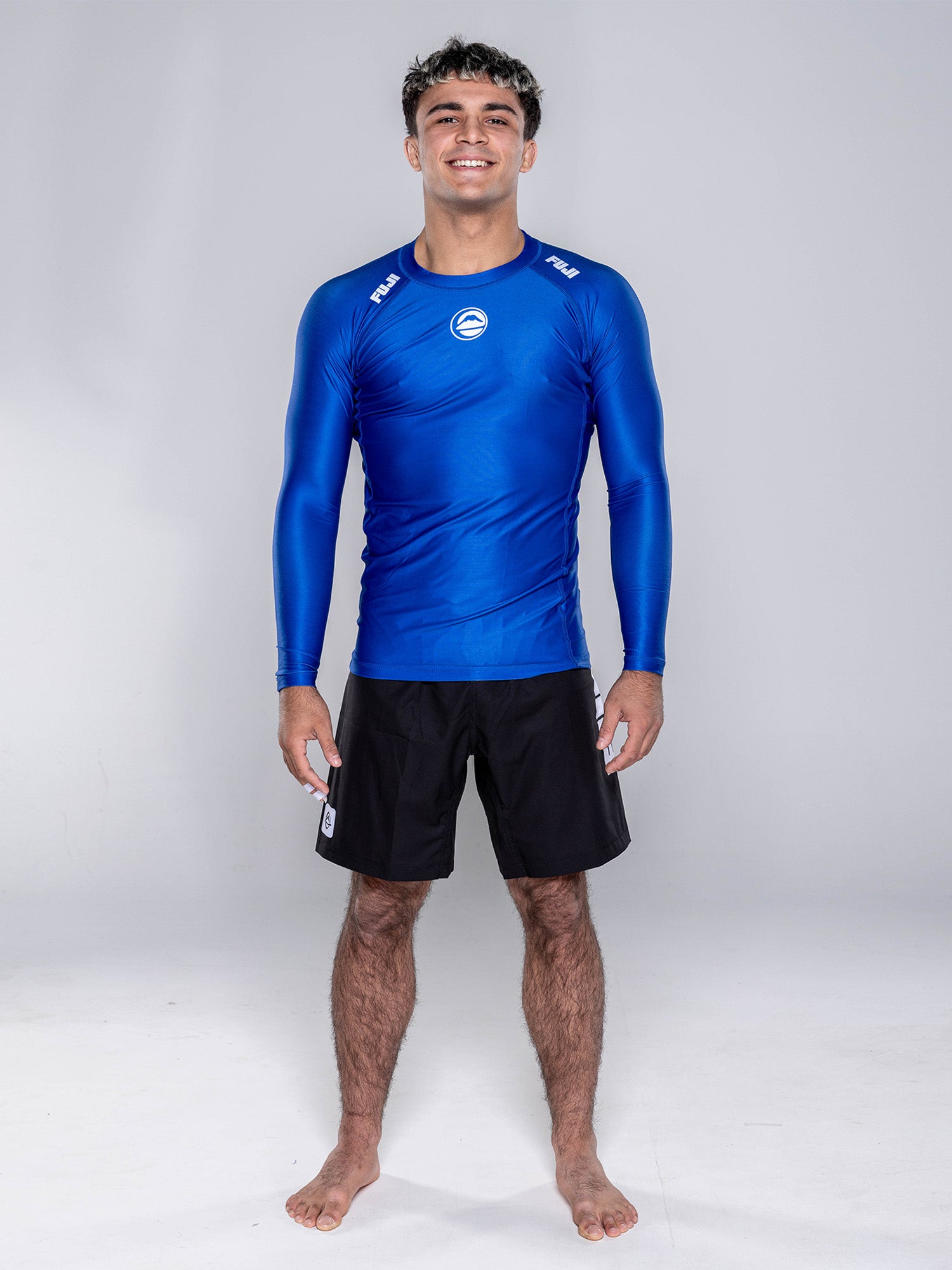 A young man stands barefoot on a white floor, smiling. He wears the Baseline 2.0 Long Sleeve Rashguard Navy and black shorts against a plain, light gray background.