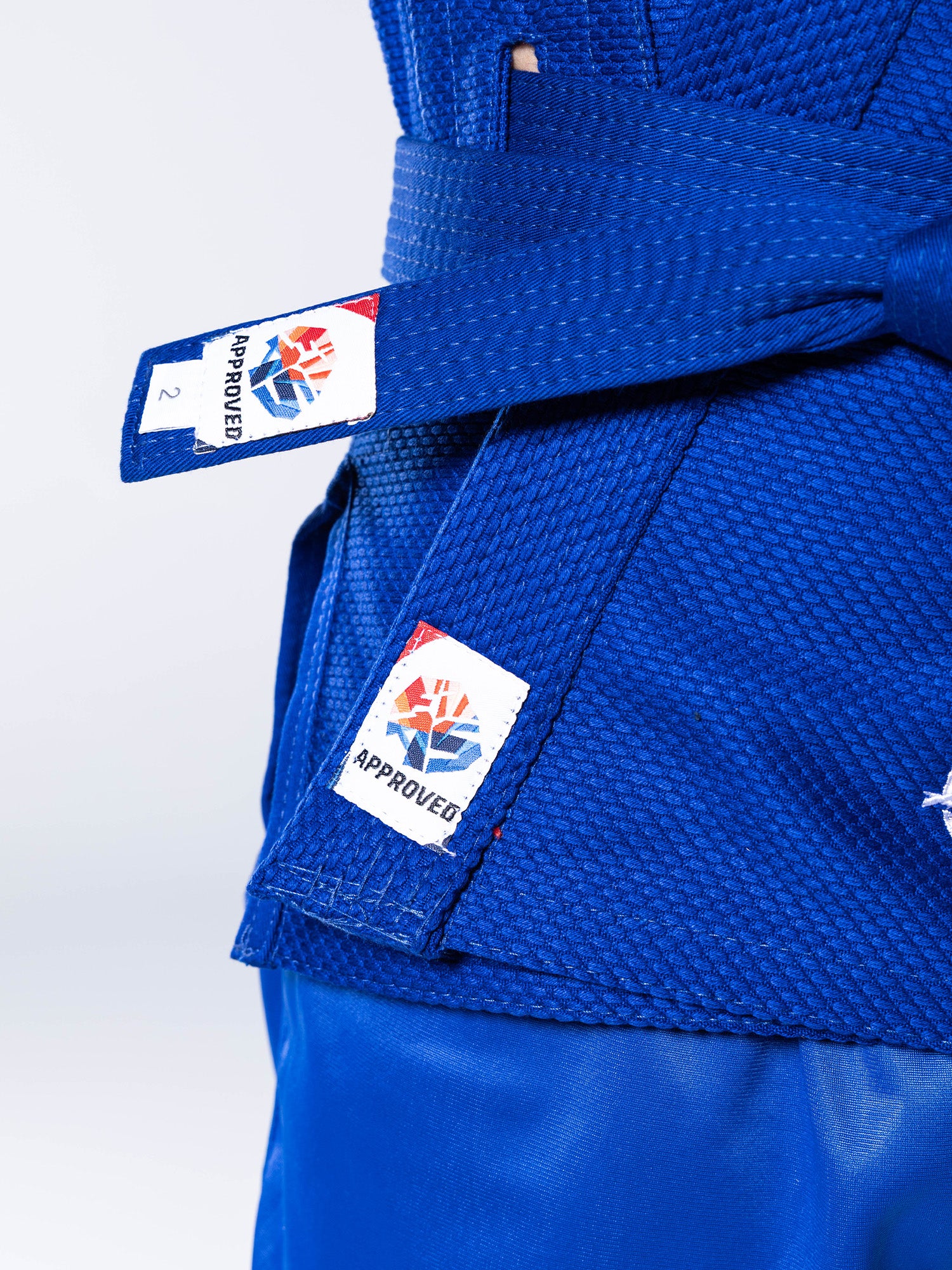 Close-up of a person wearing the FIAS Sambo Kurtka Jacket in blue with a matching blue belt tied at the front. The jacket features two tags displaying a colorful logo and the word “APPROVED.”.