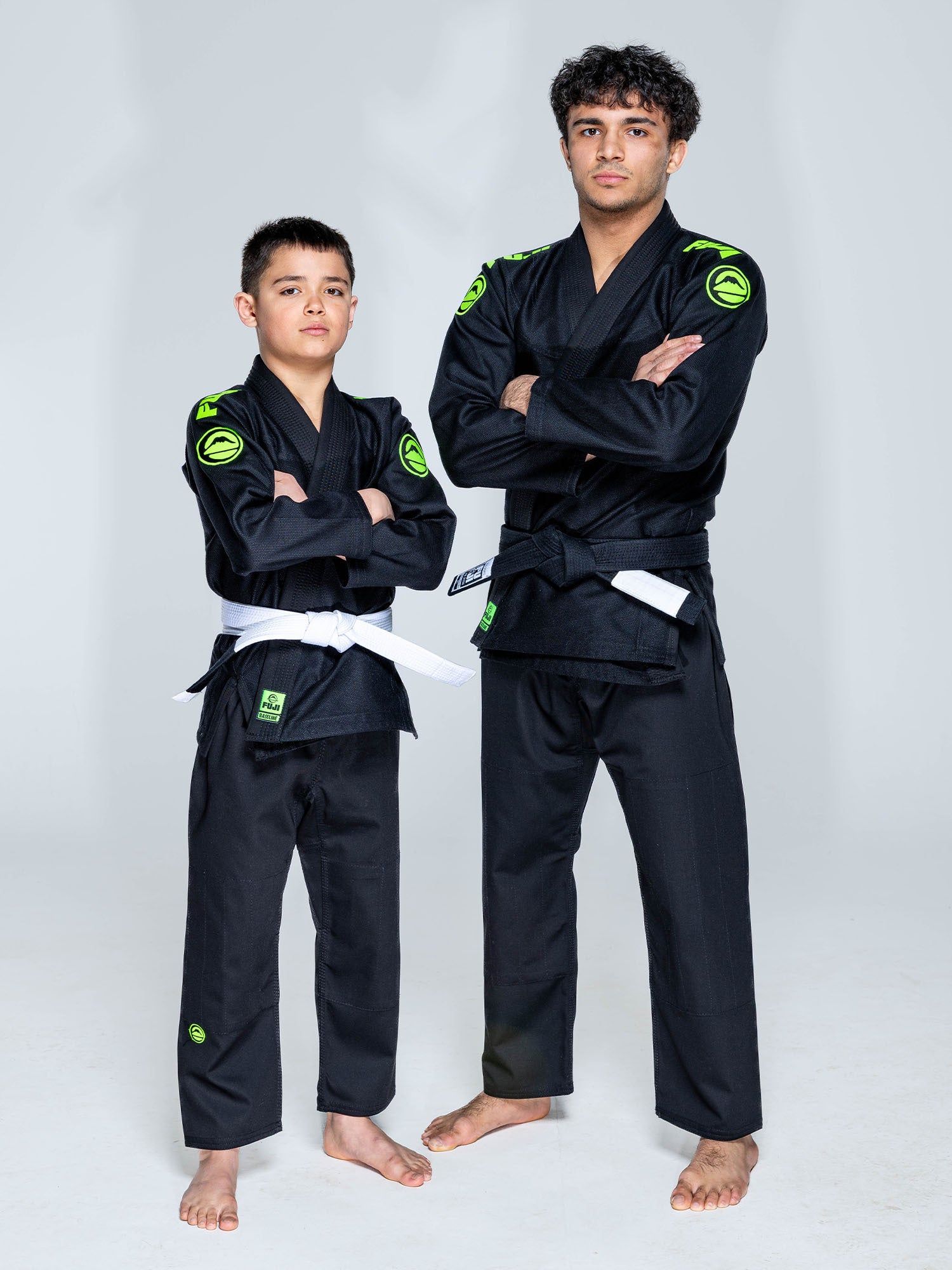 Two kids wear the Kids Baseline 2.0 Slim Fit BJJ Gi Black Green uniforms with white belts, standing barefoot side by side, arms crossed, confidently facing the camera against a plain light-colored background.