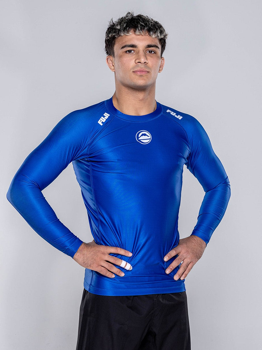 A person stands barefoot on a white floor, hands on hips, wearing the Baseline 2.0 Long Sleeve Rashguard Navy and black athletic shorts, facing the camera against a plain light gray background.