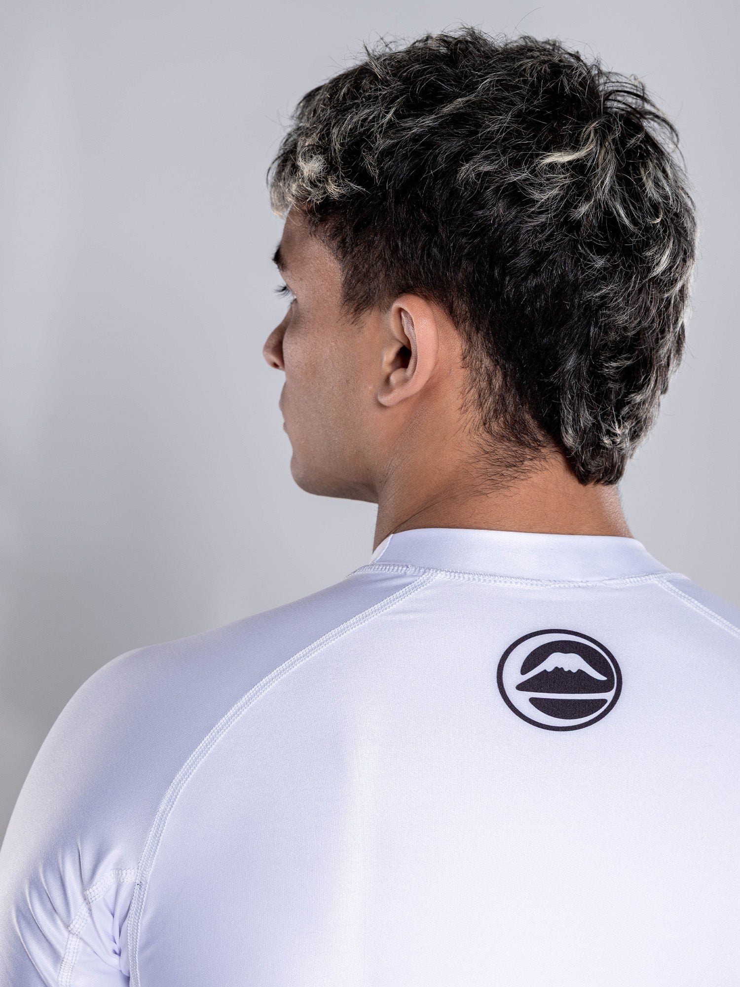 A person with short, dark hair and light highlights is seen from behind wearing the Baseline 2.0 Short Sleeve Rashguard White, which features a black circular mountain logo on the upper back against a plain light background.