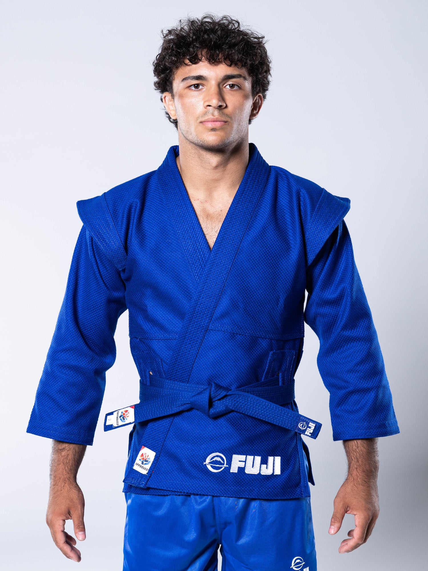 A young man with curly hair stands facing forward, wearing a blue FIAS Sambo Kurtka jacket with a matching blue belt. The jacket features FIAS branding, and he has a neutral expression against a plain background.