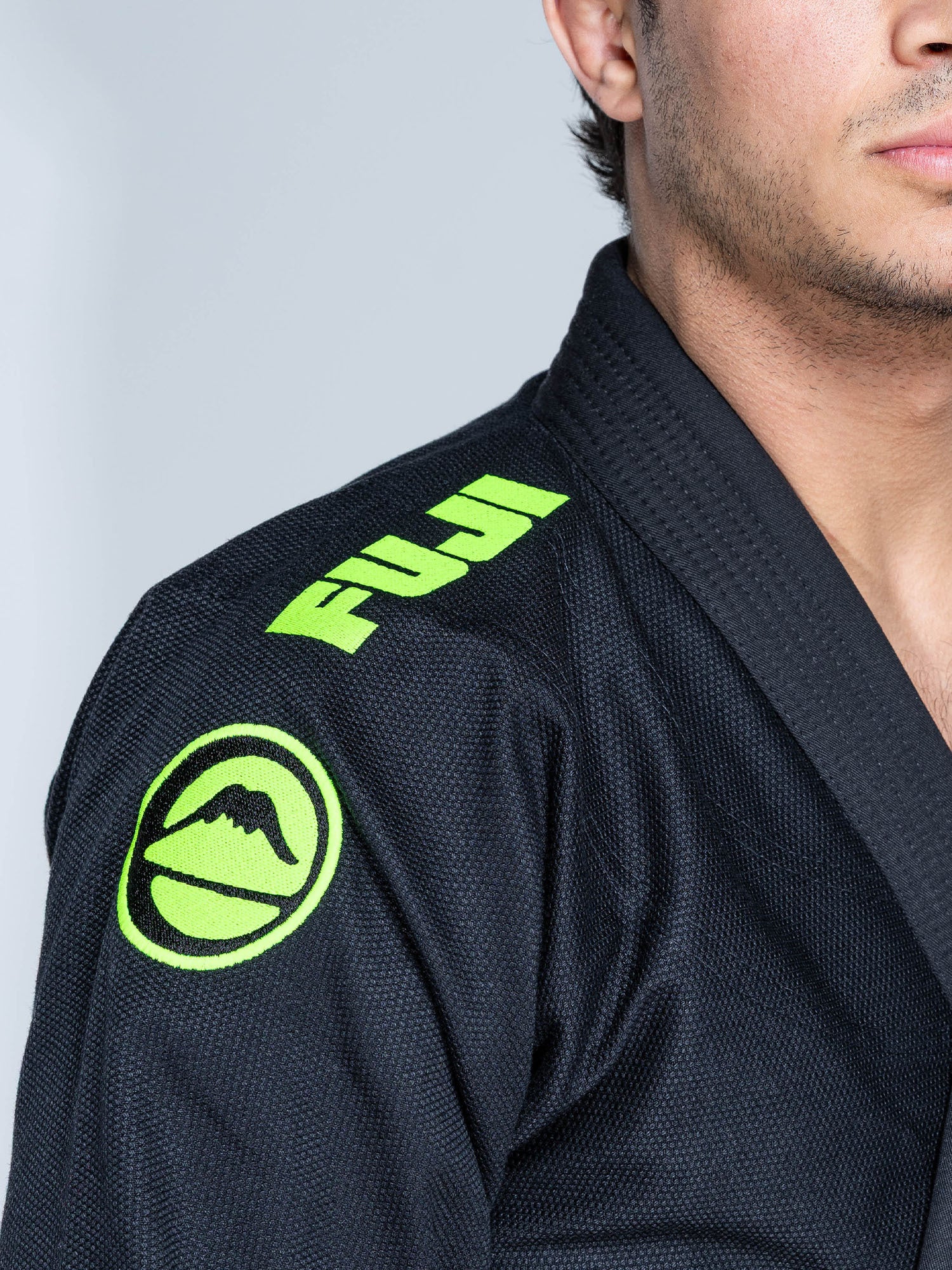 A person wears the Baseline 2.0 Slim Fit BJJ Gi Black Green, featuring a neon green FUJI logo and a mountain patch on the shoulder. Only their shoulder and lower face are visible.