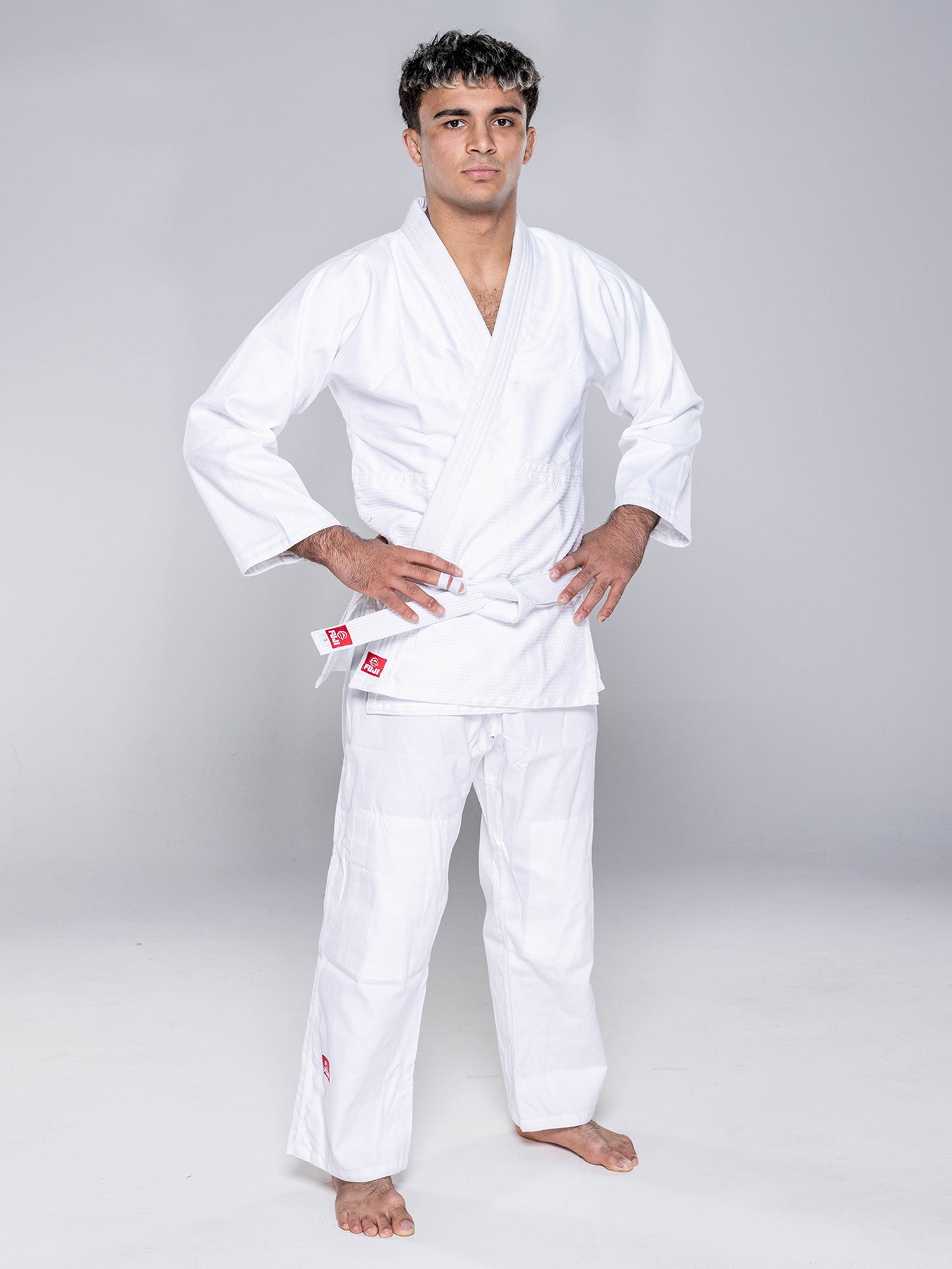 A person stands barefoot on a white floor, wearing the Essential Judo Kimono White with a white belt, hands on hips, and looking forward against a plain light gray background.