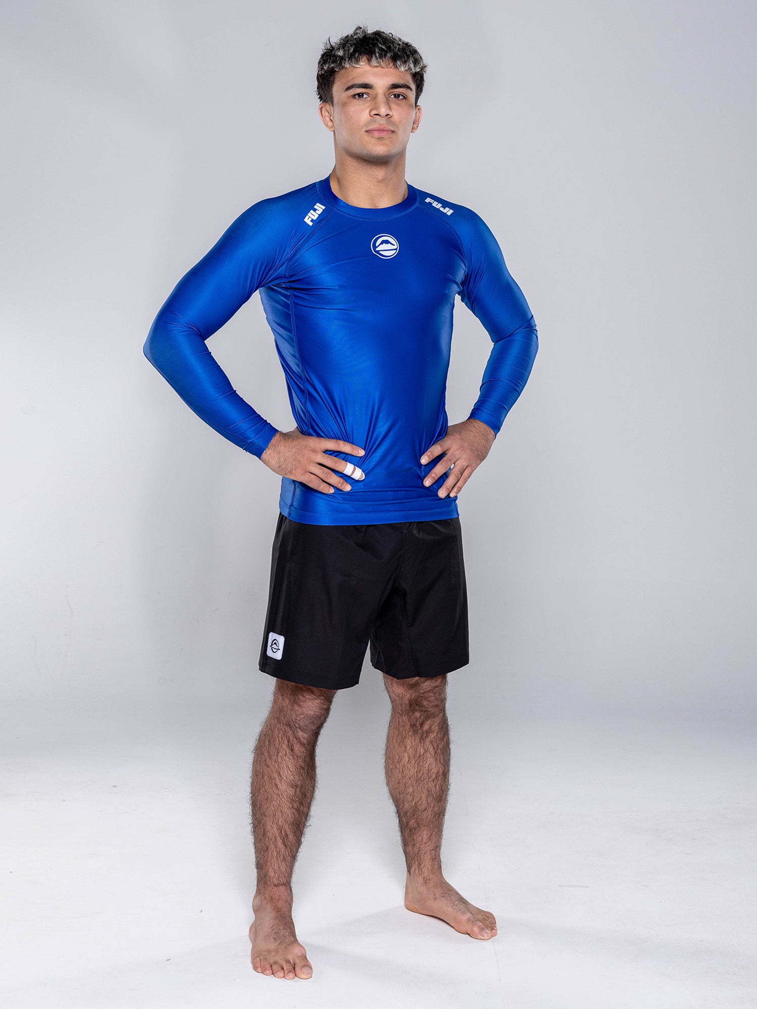 A young man stands barefoot on a white background, confidently wearing the Baseline 2.0 Long Sleeve Rashguard Navy and black shorts with his hands on his hips.