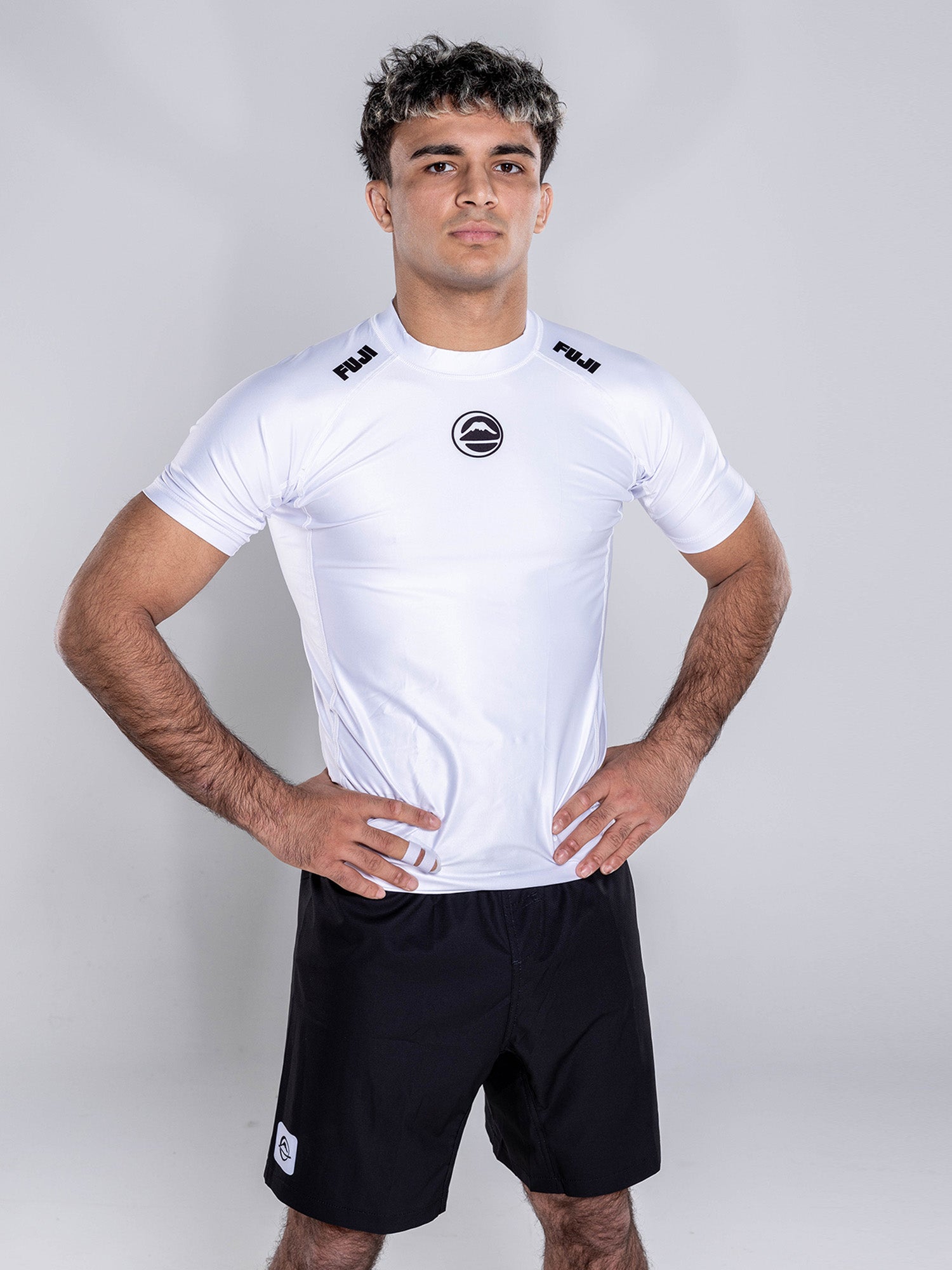 A man with short curly hair stands confidently with hands on hips, wearing the Baseline 2.0 Short Sleeve Rashguard White and black shorts against a plain light background.