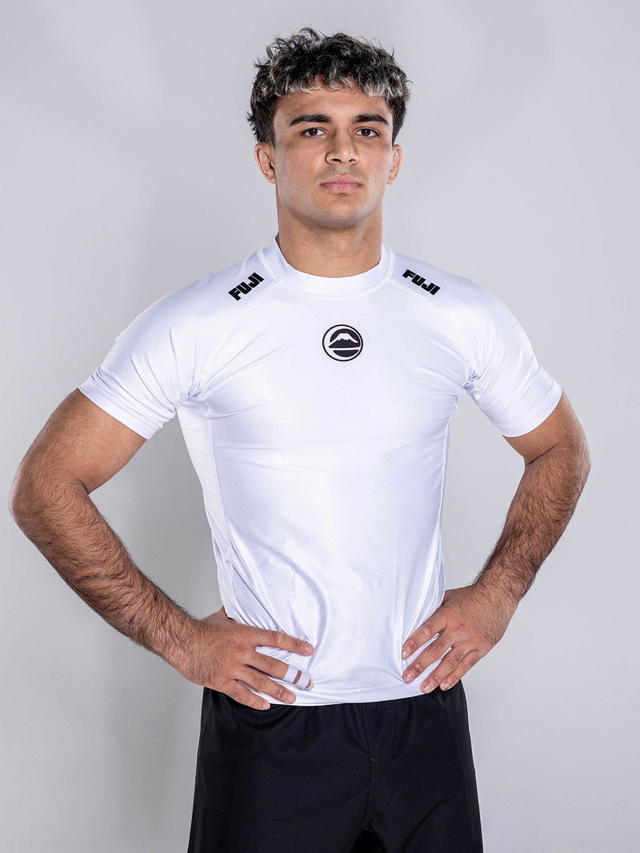 A man with short curly hair stands confidently with hands on hips, wearing the Baseline 2.0 Short Sleeve Rashguard White and black shorts against a plain light background.