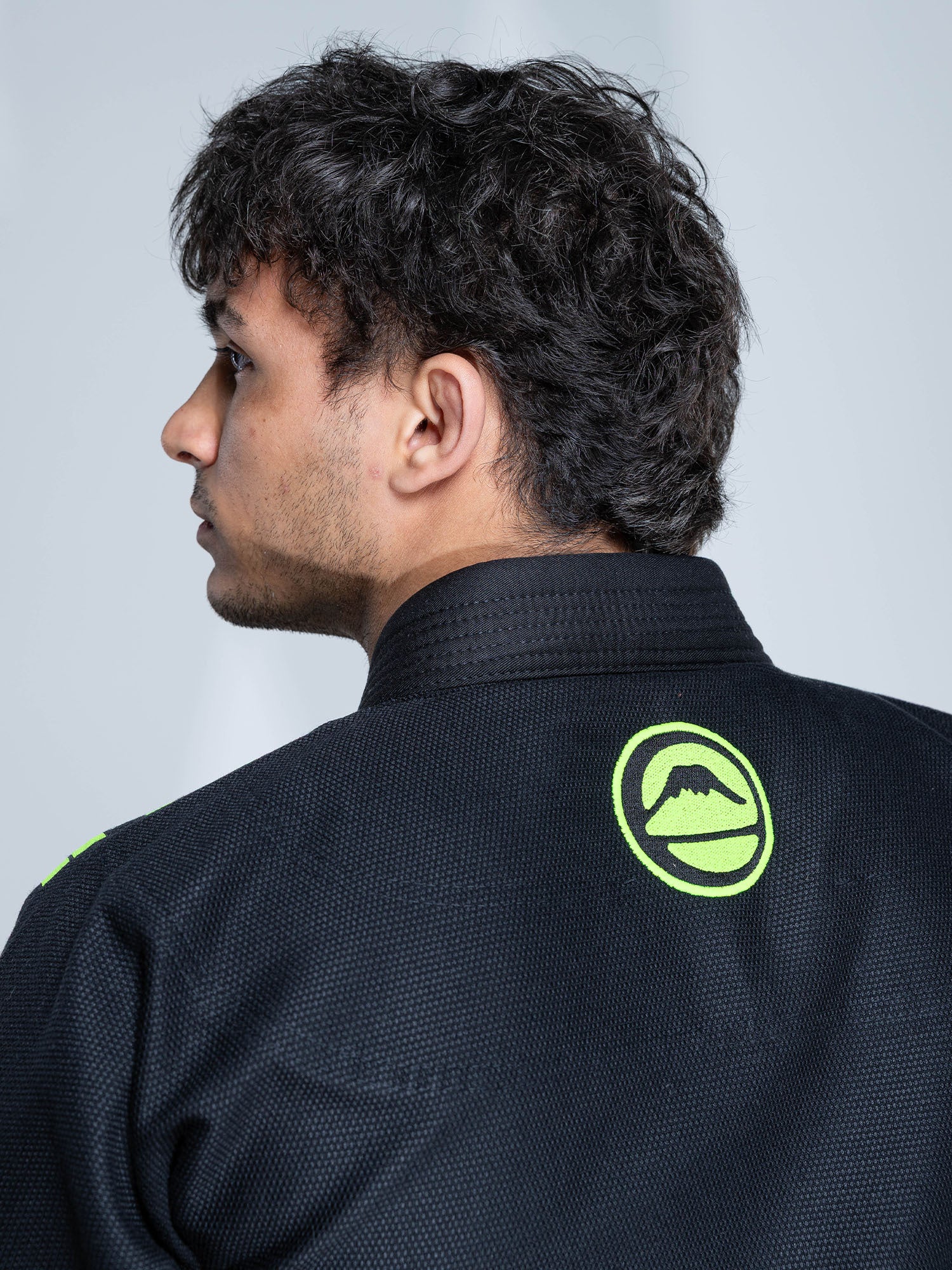 A person with short, curly dark hair is seen from behind wearing the Baseline 2.0 Slim Fit BJJ Gi Black Green, which features a green mountain logo patch on the back. The background is plain and light-colored.