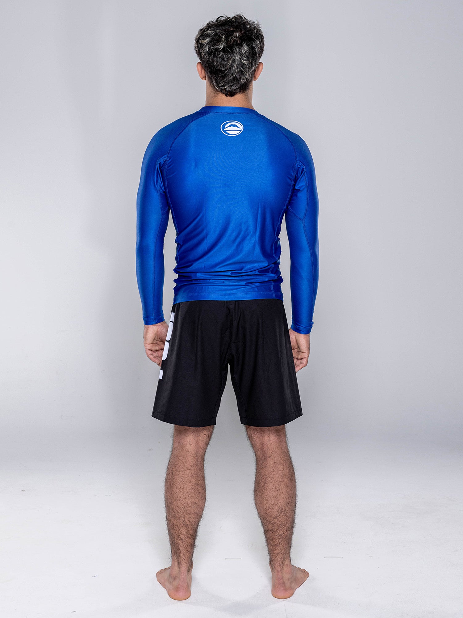 A man stands with his back to the camera, wearing the Baseline 2.0 Long Sleeve Rashguard in navy and black shorts, set against a plain light gray background.
