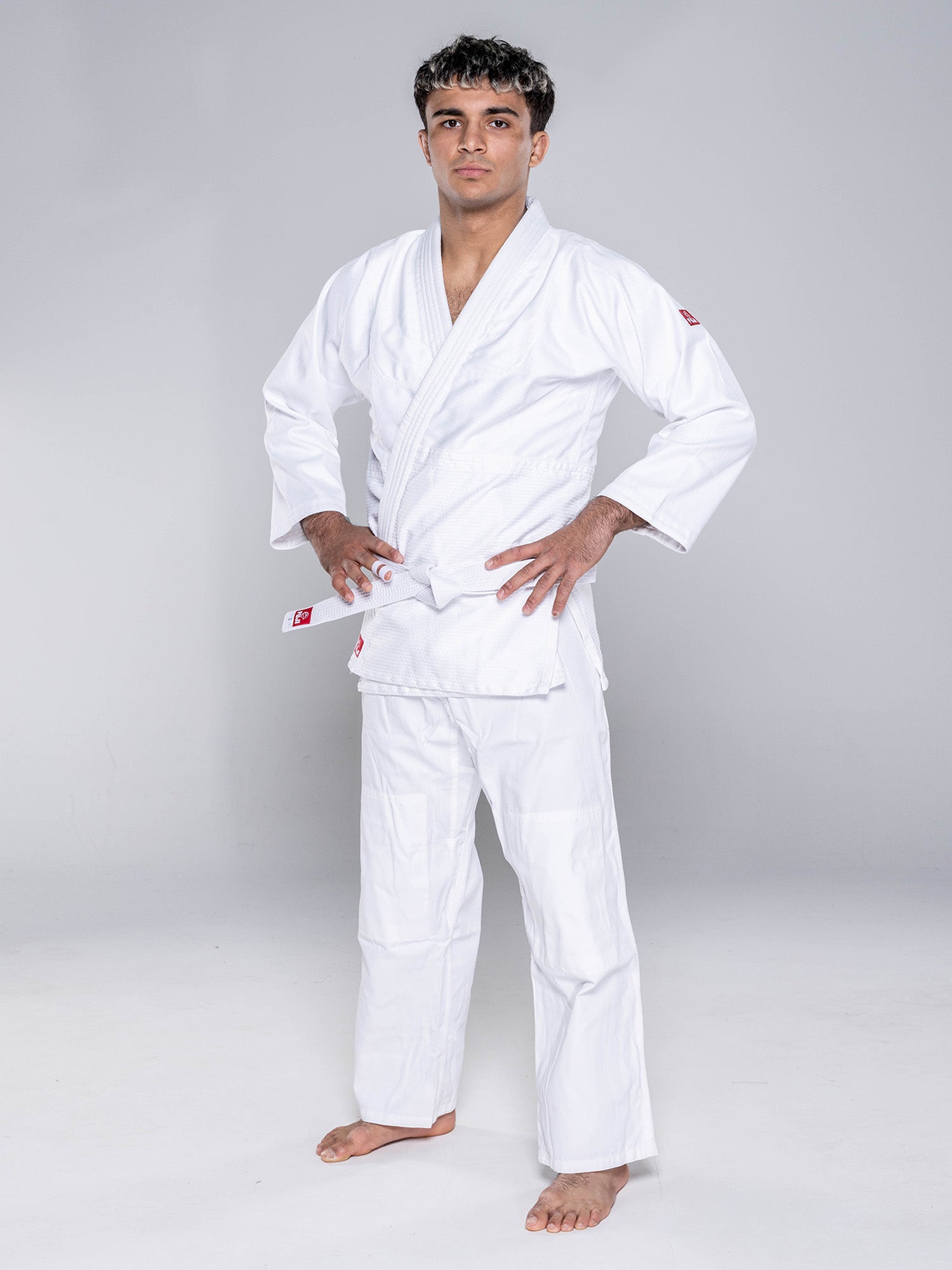 A person stands barefoot on a white floor, wearing the Essential Judo Kimono White with a belt, hands on hips, looking confidently at the camera against a plain light gray background.