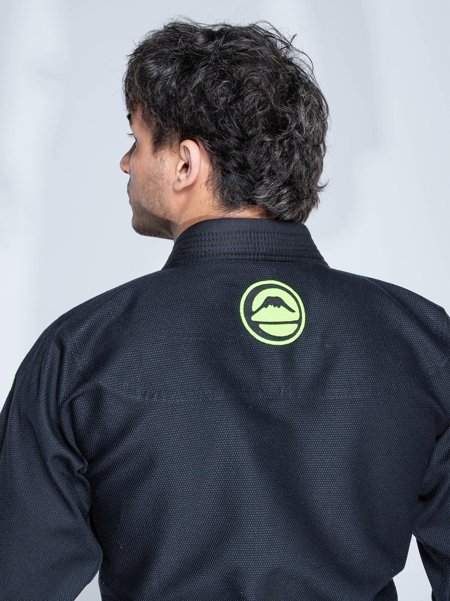 A person wearing the Baseline 2.0 Slim Fit BJJ Gi Black Green, featuring a green circular mountain logo on the back, is shown from behind against a plain light background.
