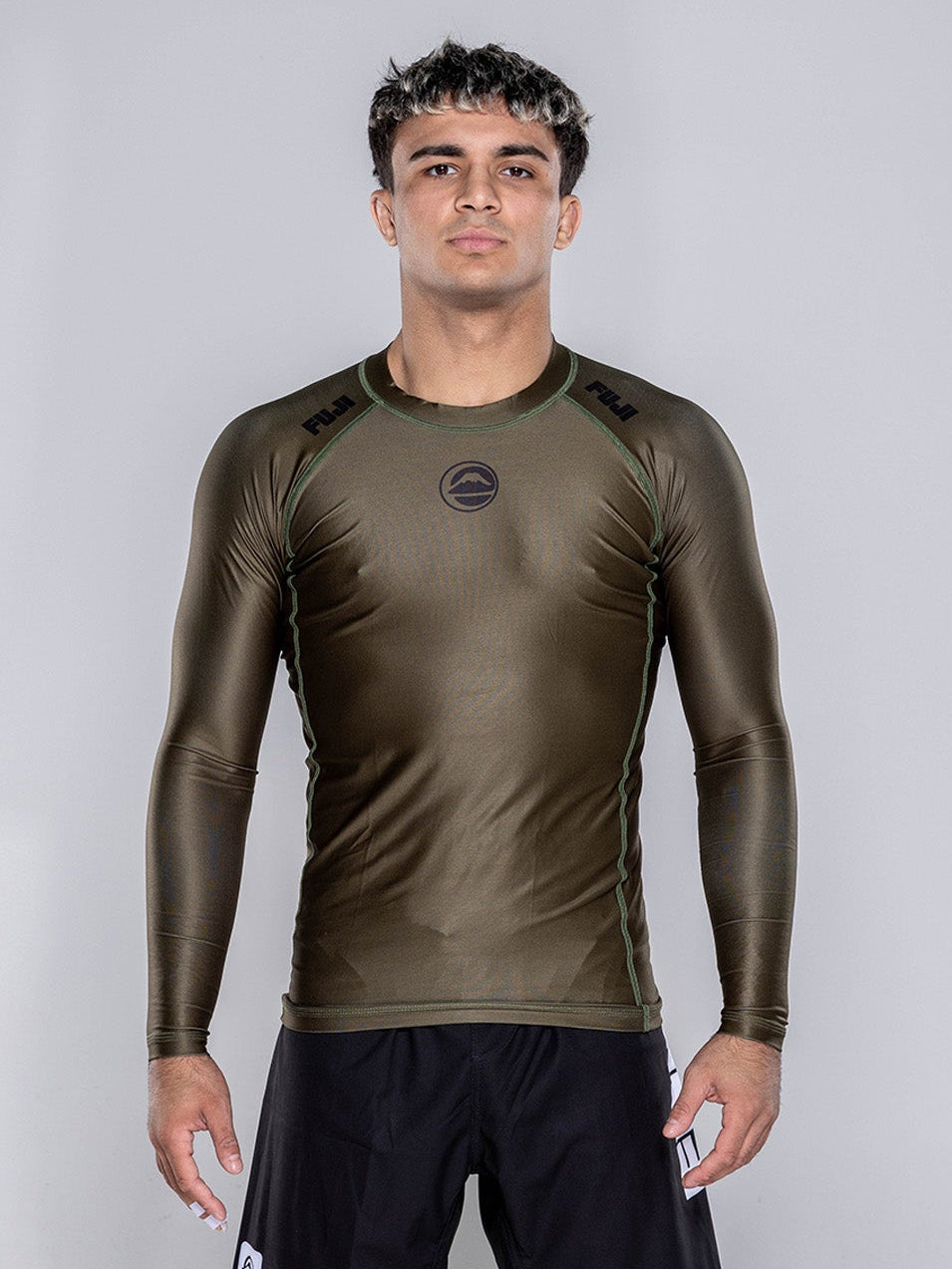 A man stands barefoot on a white floor, wearing the Baseline 2.0 Long Sleeve Rashguard in Military Green and black athletic shorts, facing forward with arms relaxed by his sides against a plain light gray background.