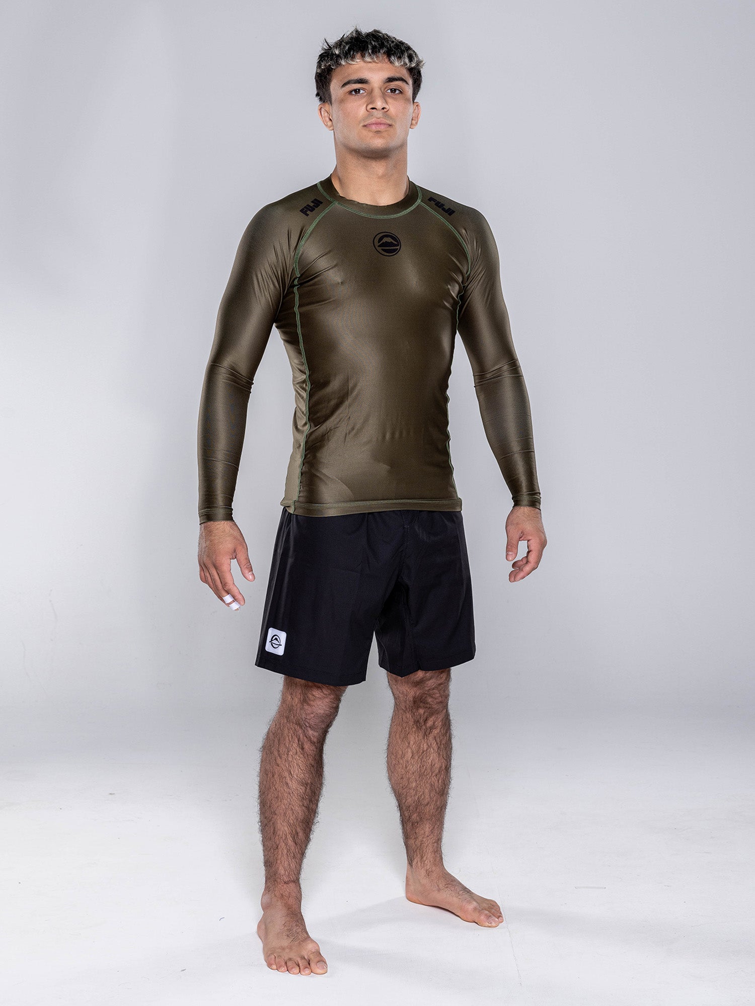 A person stands barefoot on a white floor, wearing the Baseline 2.0 Long Sleeve Rashguard in Military Green and black shorts. The background is plain light gray, and the person faces forward with a neutral expression.