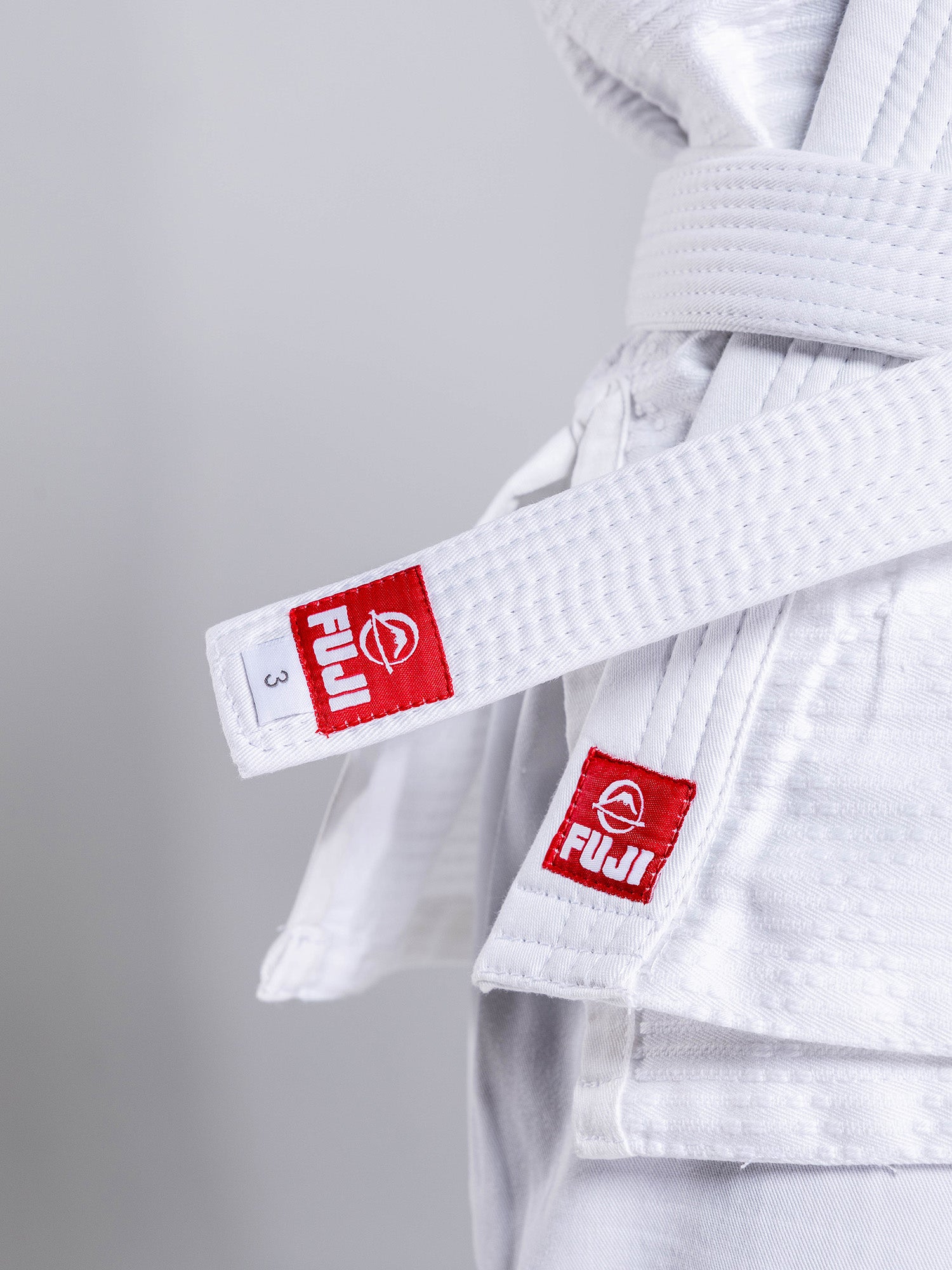 A close-up of a person wearing the Essential Judo Kimono White with a matching white belt featuring red FUJI brand labels, both neatly tied against a plain light gray background.