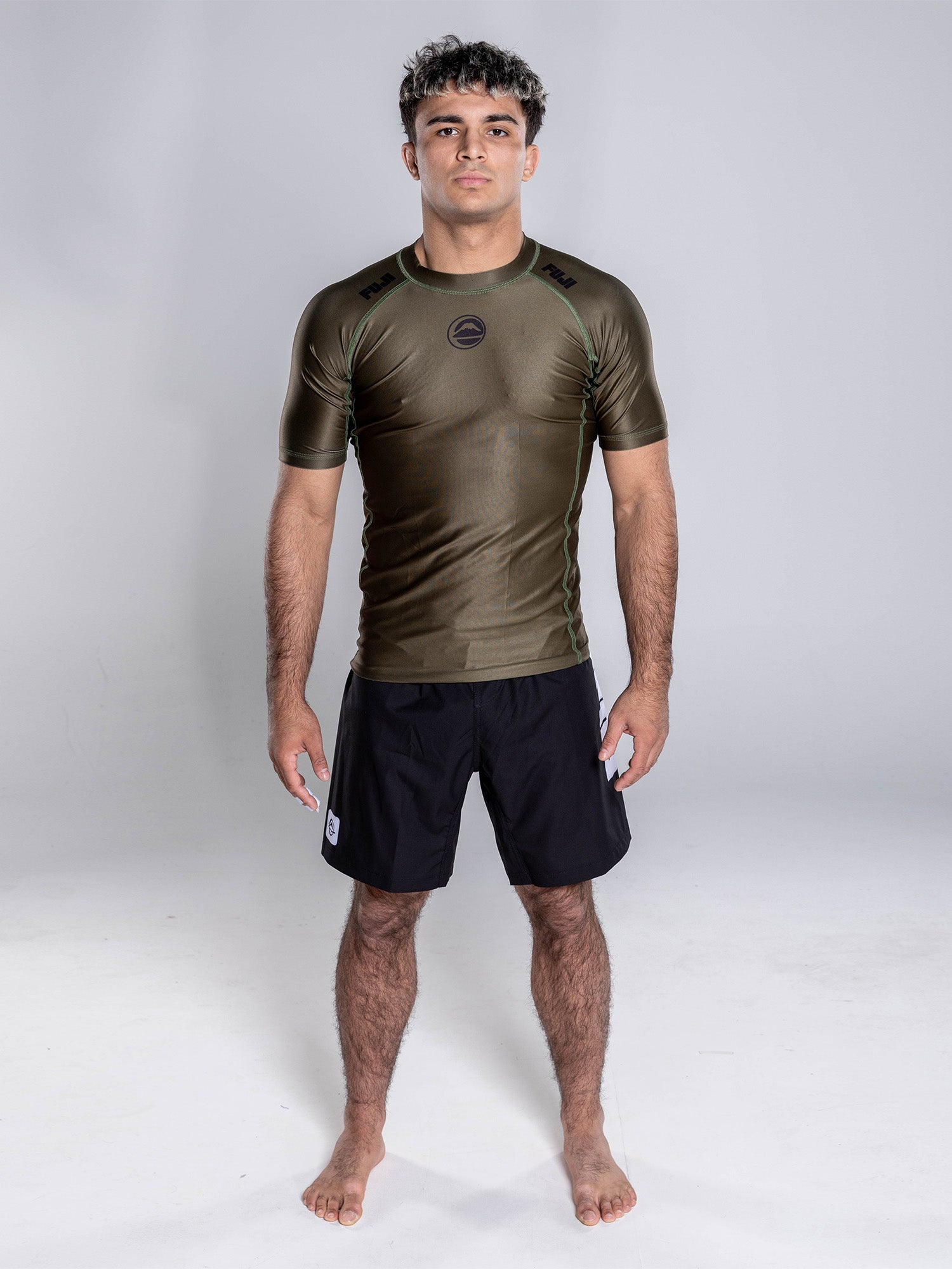 A man stands barefoot on a white background, wearing the Baseline 2.0 Short Sleeve Rashguard in Military Green and black shorts, facing forward with his arms relaxed at his sides.