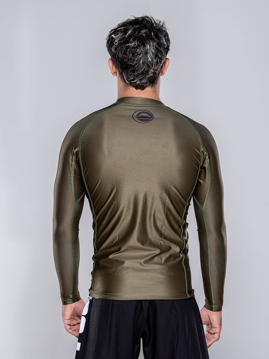 A man stands barefoot on a white floor, facing away from the camera. He is wearing the Baseline 2.0 Long Sleeve Rashguard in Military Green and black shorts, with a plain light gray background.