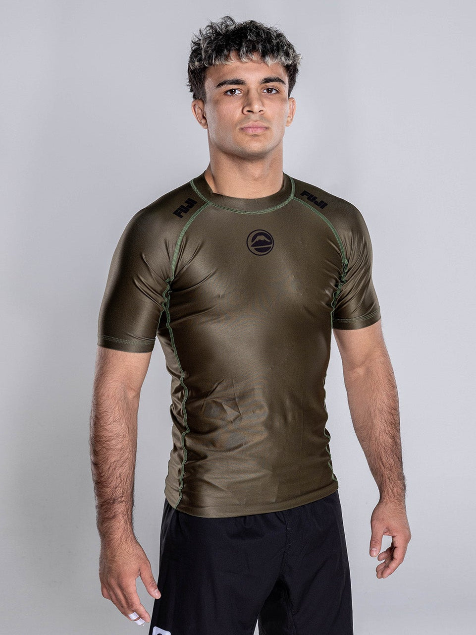A man stands barefoot on a white studio floor wearing the Baseline 2.0 Short Sleeve Rashguard in Military Green and black athletic shorts, looking directly at the camera with a neutral expression.