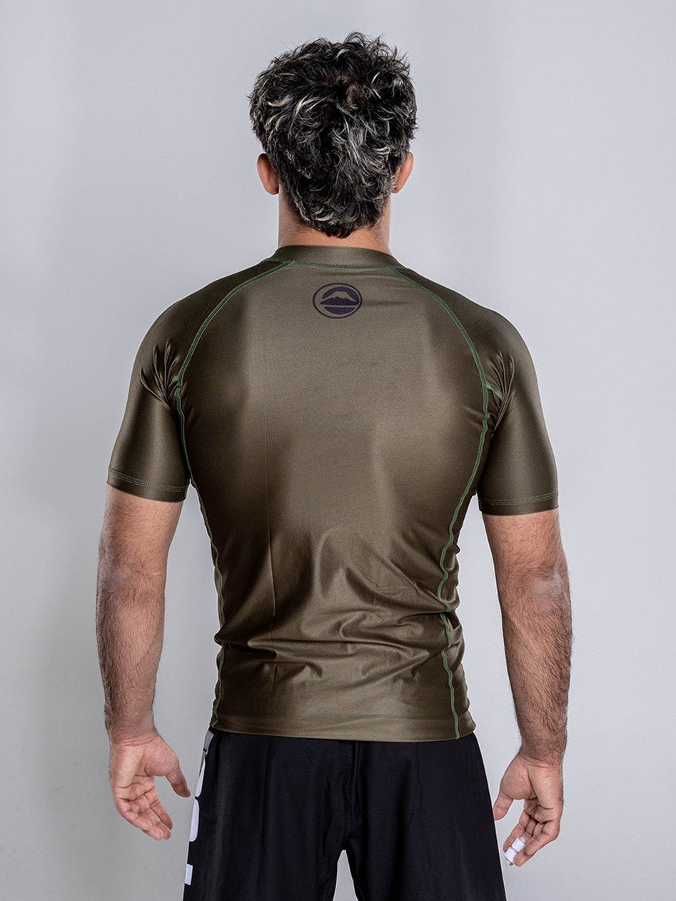 A man stands barefoot on a white background, facing away from the camera, wearing the Baseline 2.0 Short Sleeve Rashguard in Military Green and black shorts with his arms relaxed by his sides.